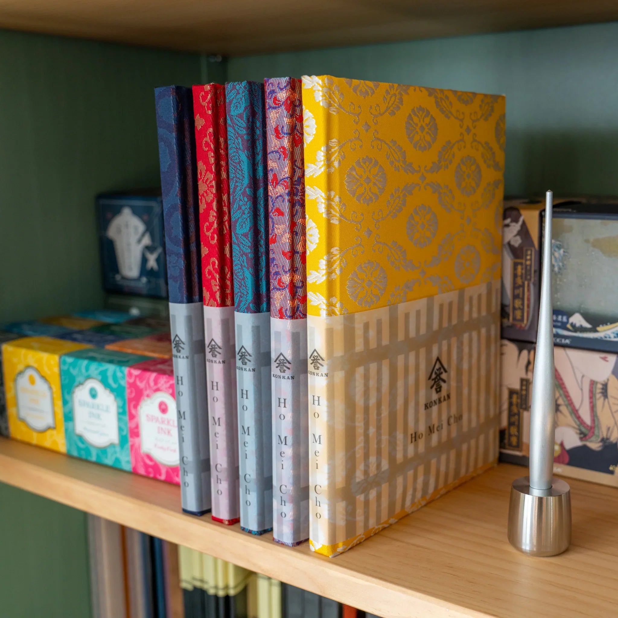 Ko Aoi Gold Nishijin silk Homeicho notebook with KONKAN Ho Mei Cho band on shelf beside other colorway notebooks