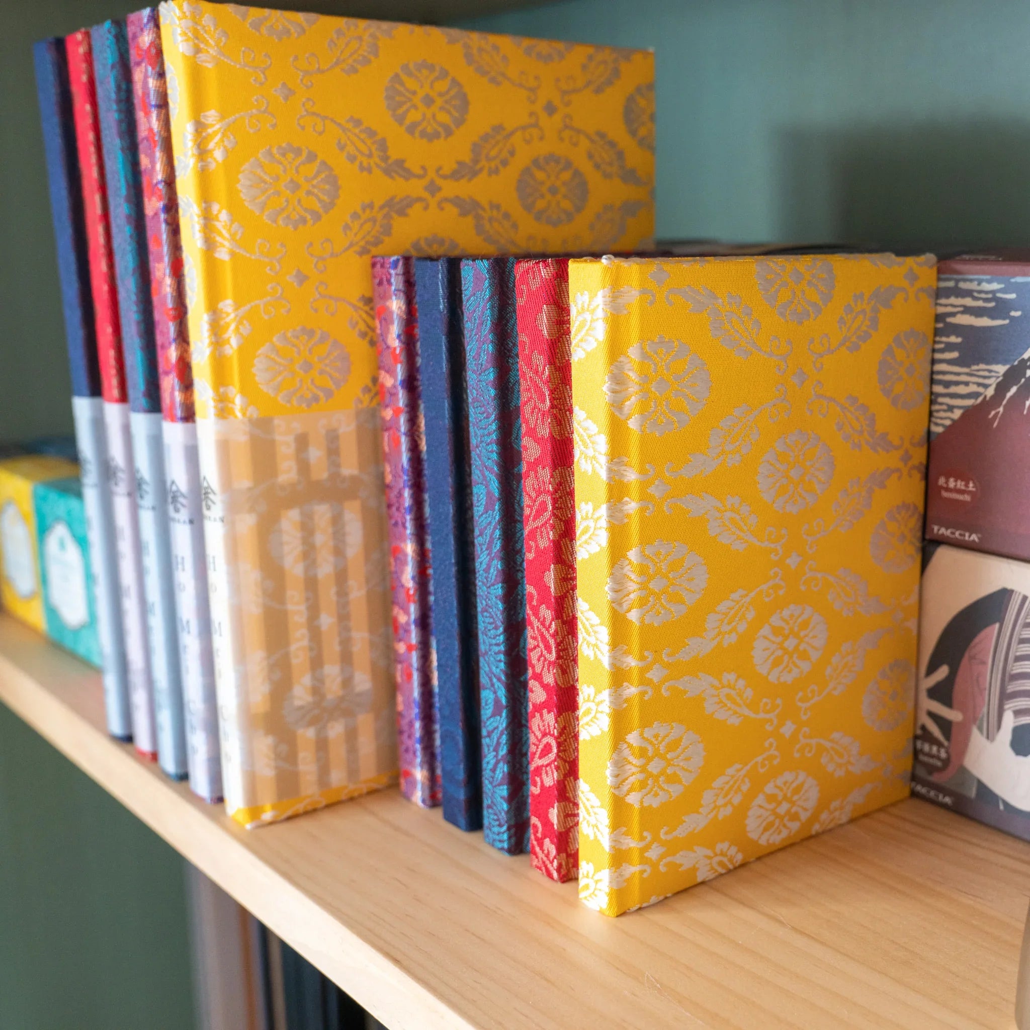 Ko Aoi Gold Nishijin silk Homeicho notebook standing on wooden shelf beside other colorways including navy and red
