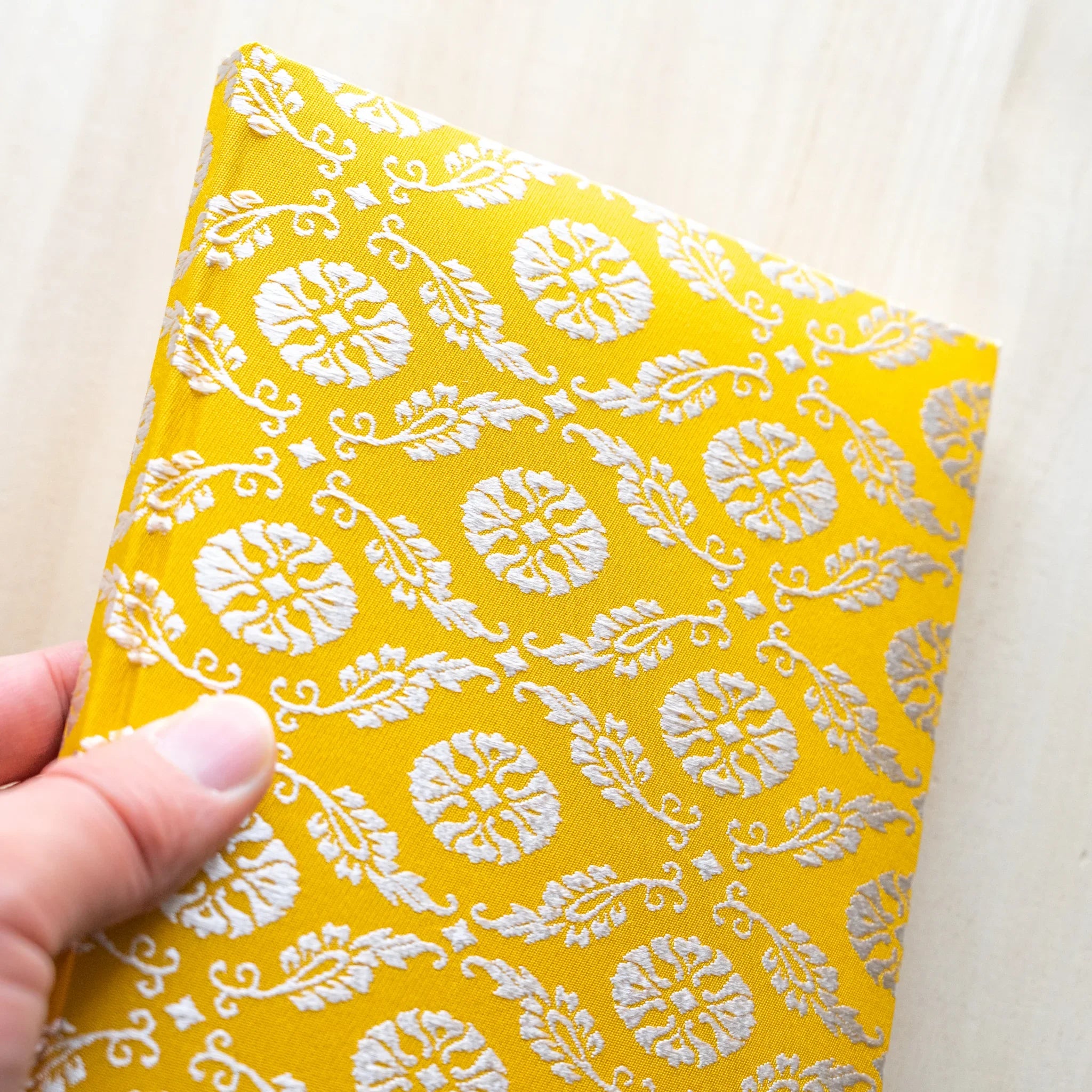 Hand holding Ko Aoi Nishijin silk Homeicho notebook in tilted vertical view, full golden Ko Aoi floral cover visible