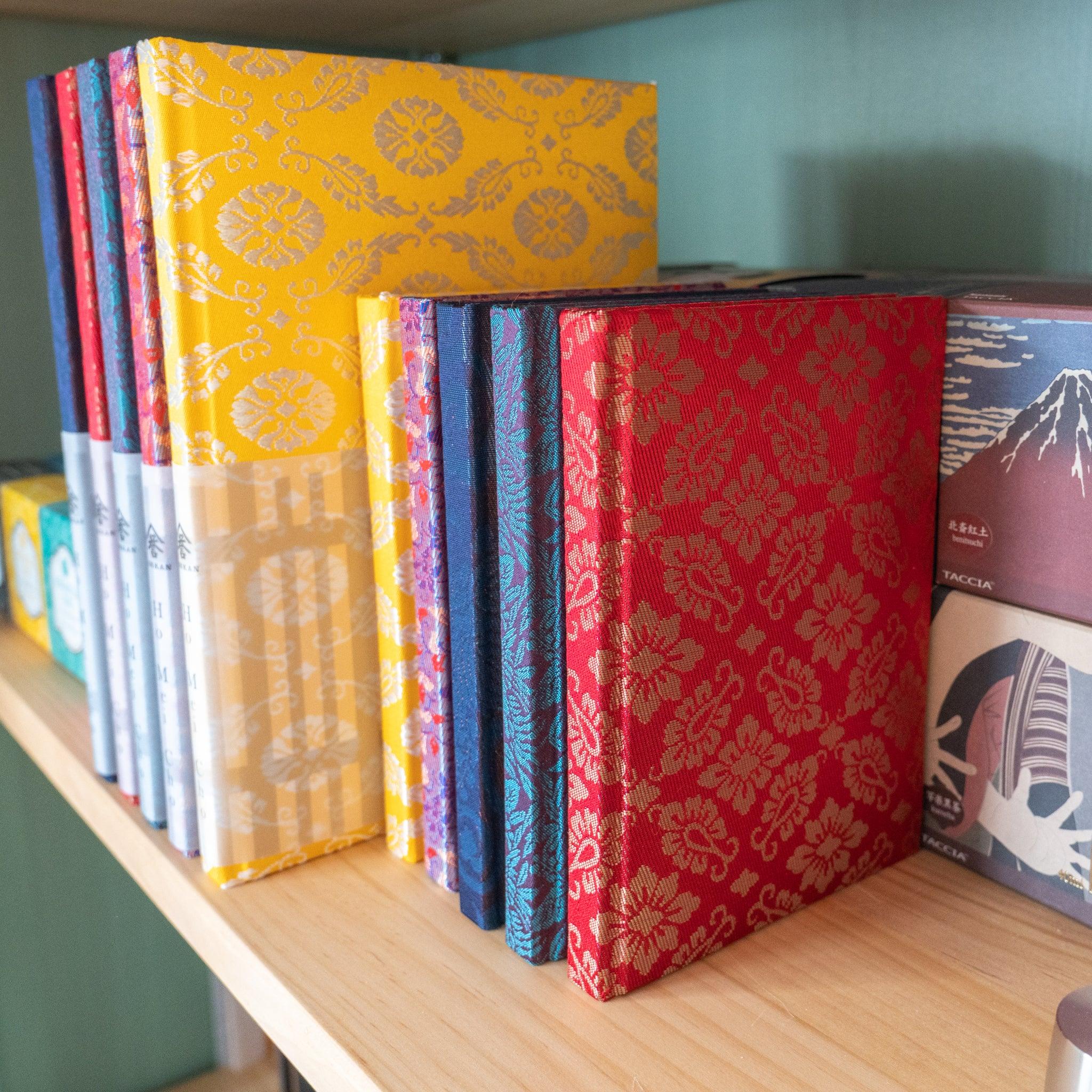 Row of Nishijin silk Homeicho notebooks standing on wooden shelf showing colorways including yellow, red, navy, and plum
