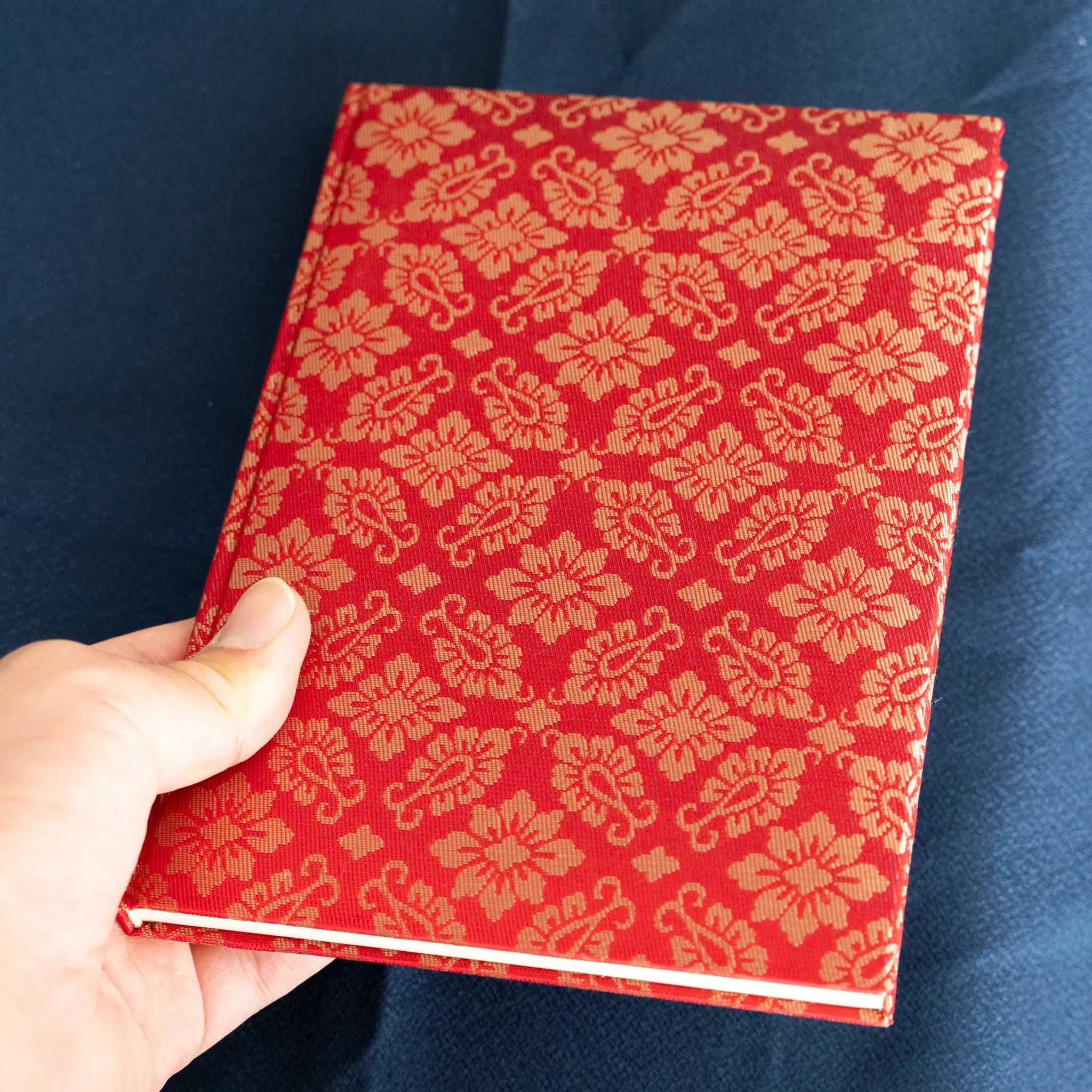 Hand holding Ko Aoi Nishijin silk Homeicho notebook in tilted vertical view, full vermilion Ko Aoi floral cover visible