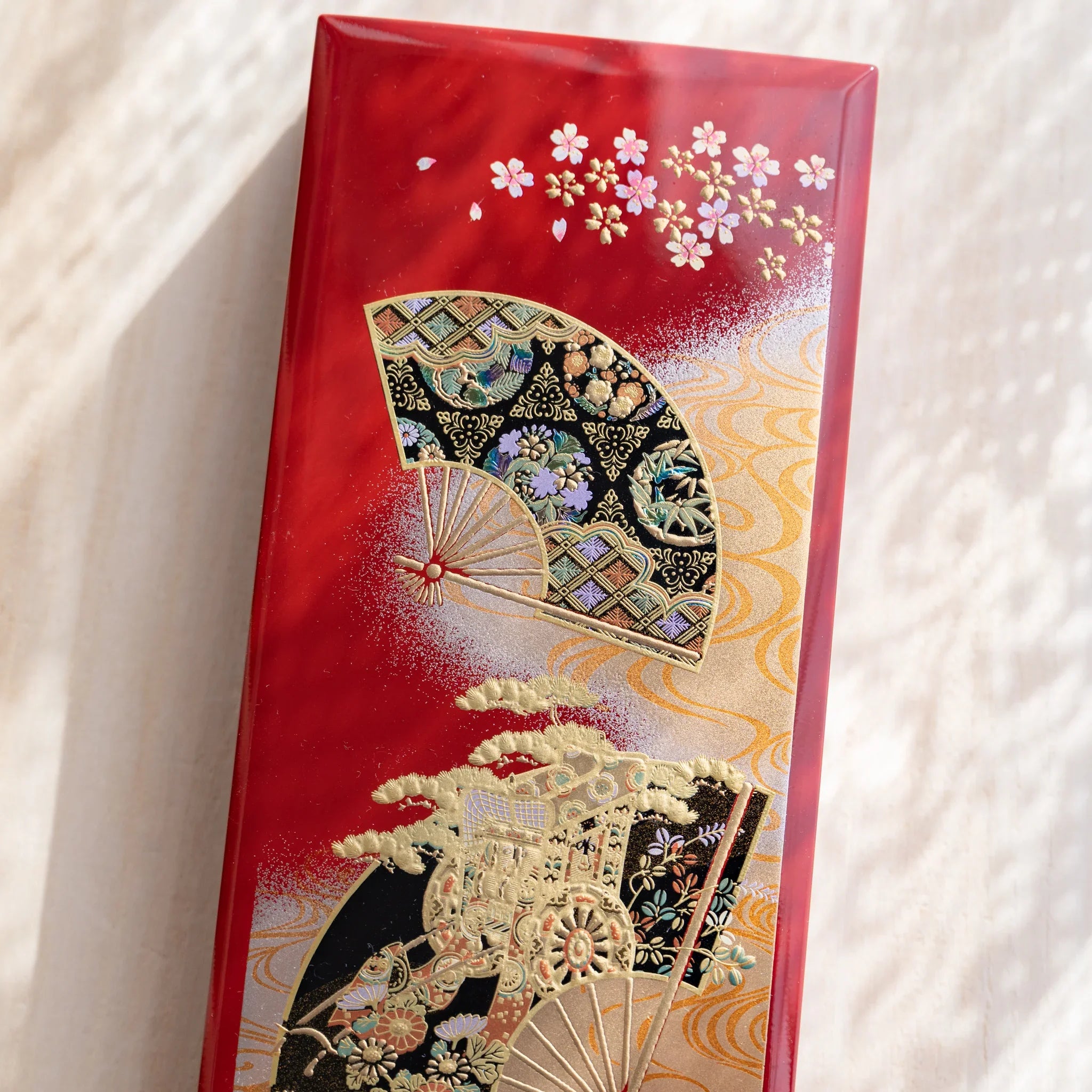 Japanese pen case closed with lid adorned by intricate gold and multicolor fan and cherry blossom motifs on red lacquer