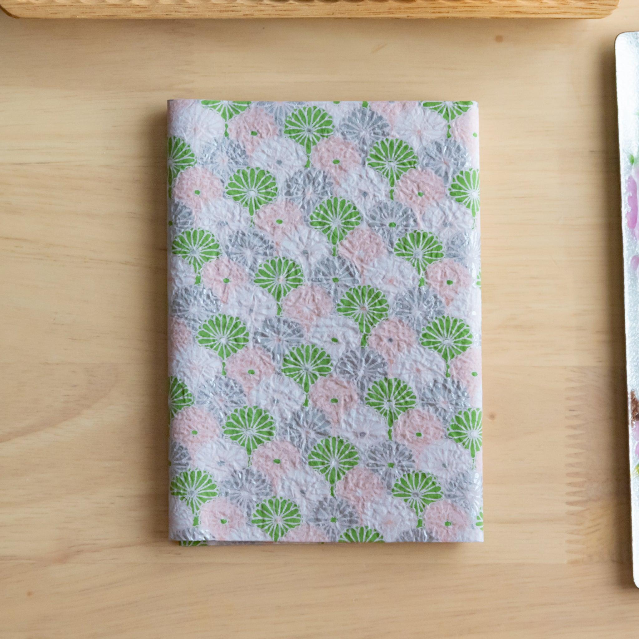 A notebook with a pink and green patterned cover and a matching gold pen, neatly placed on a wooden tray on a light wooden surface.