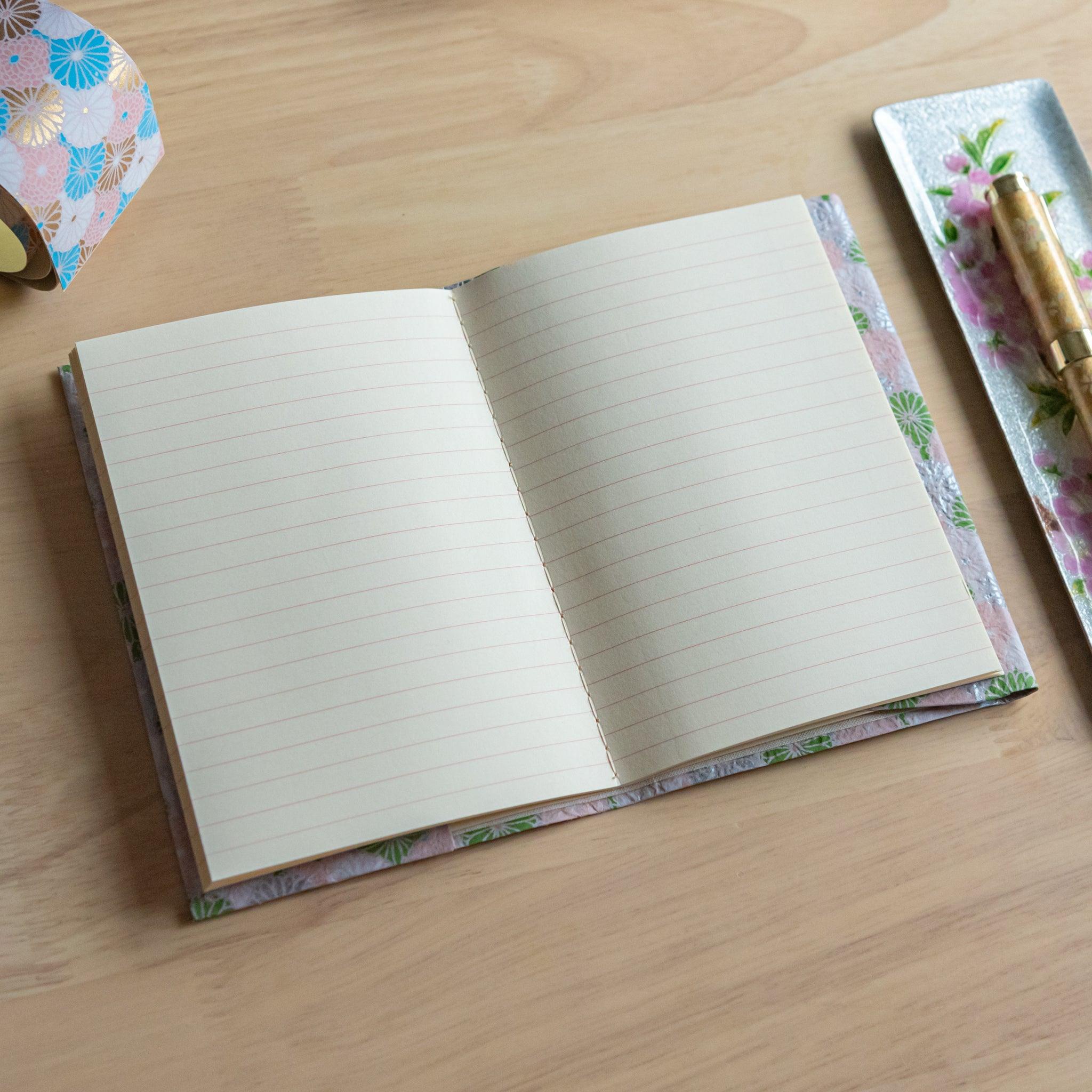 An open notebook with a pink and green patterned cover, showing lined pages, placed next to a pen on a light wooden surface.