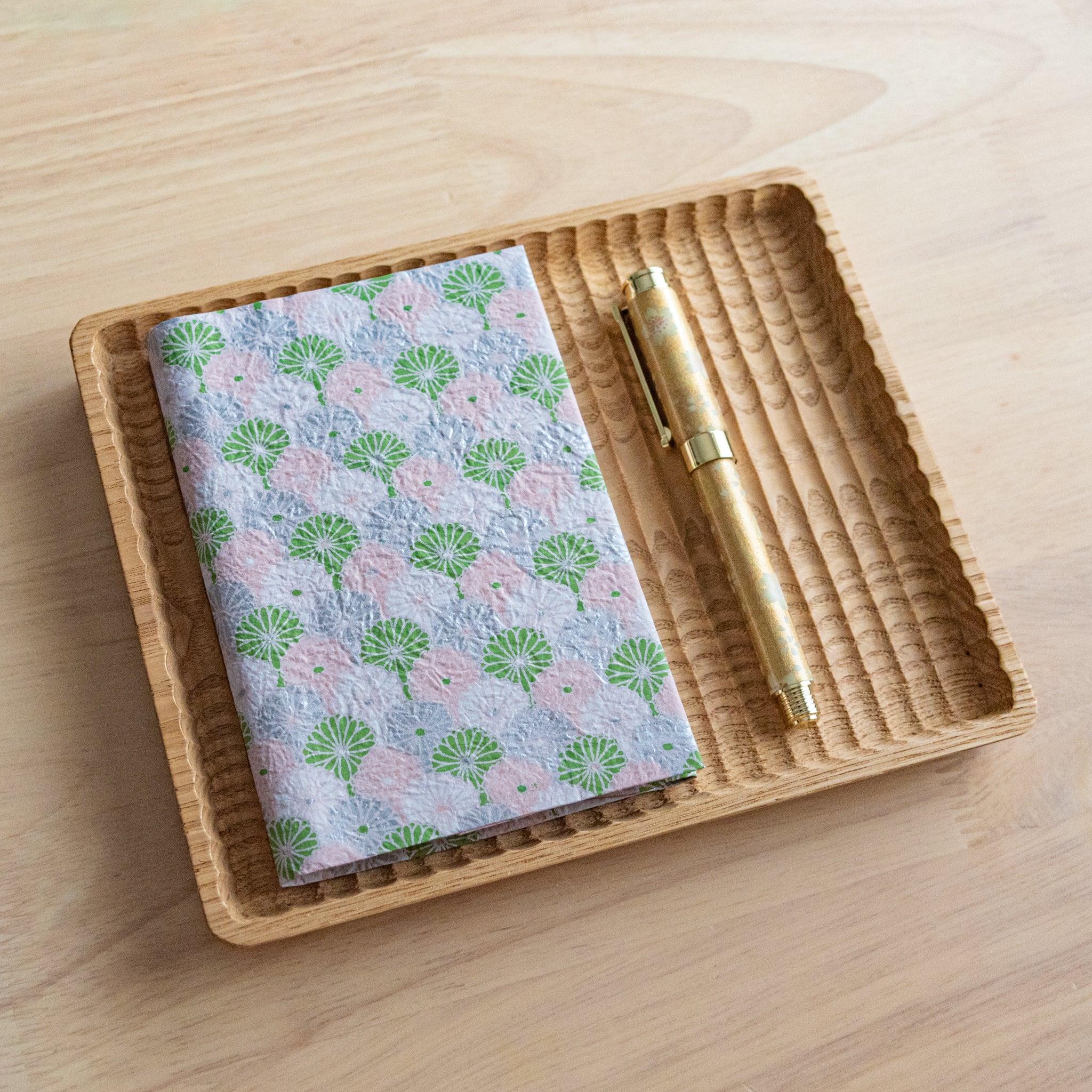 A notebook with a pink and green patterned cover, featuring intricate fan-like designs, placed on a light wooden surface.