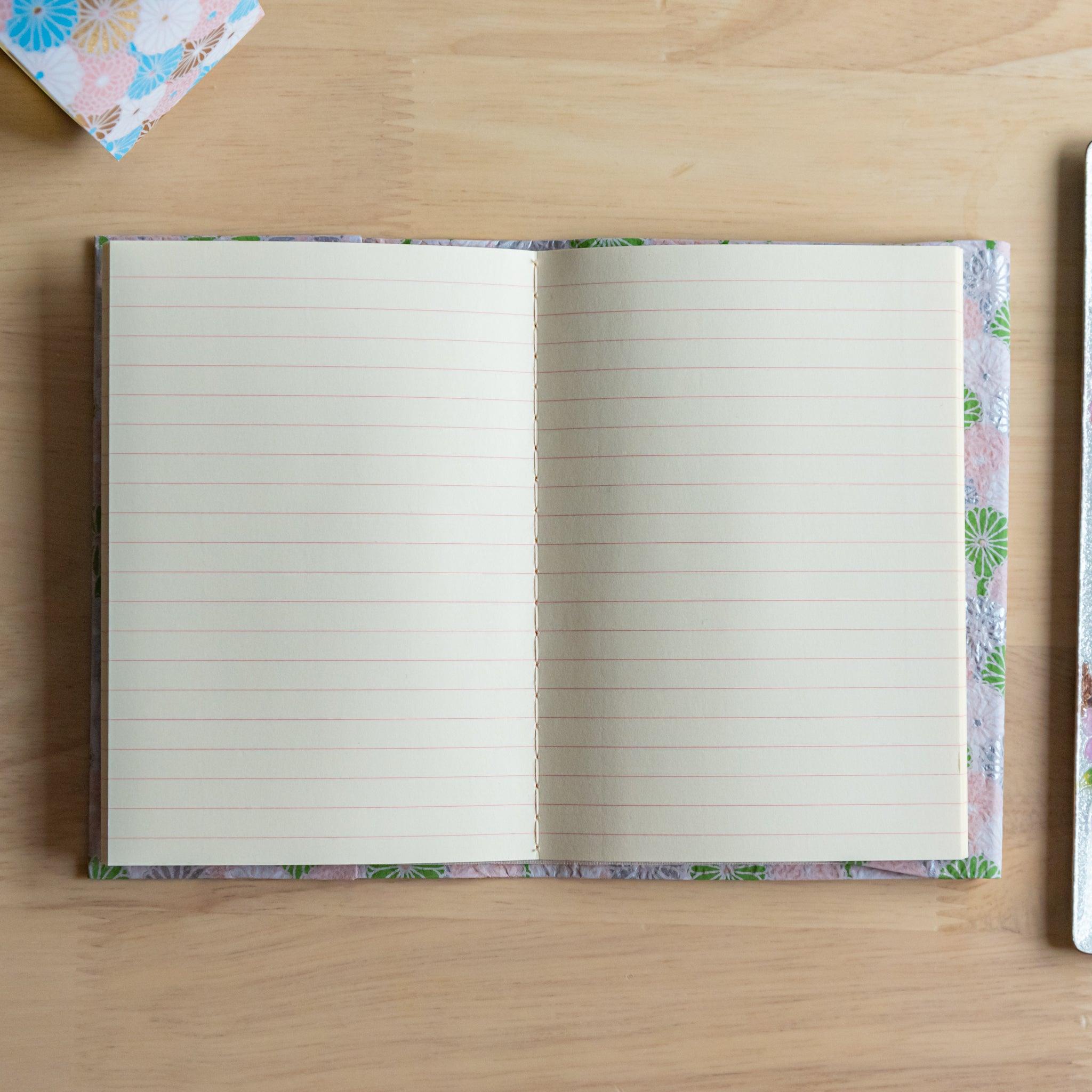 An open notebook with a pink and green patterned cover, fully revealing two lined pages, placed on a light wooden surface.