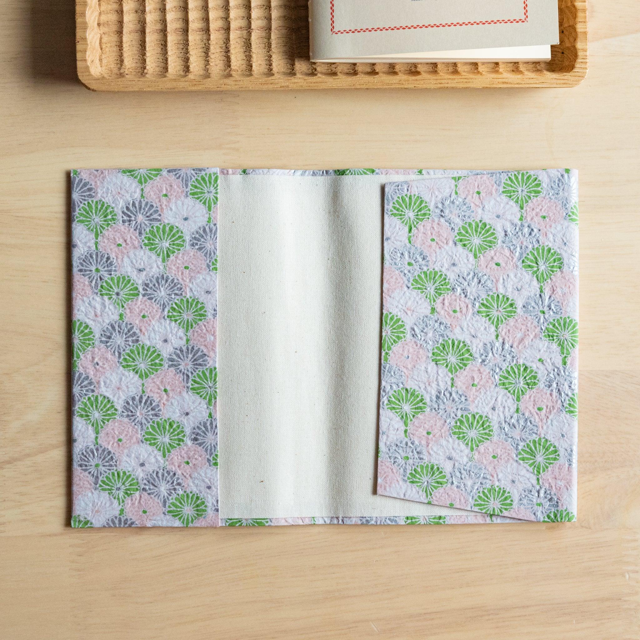 An open notebook cover with a pink and green fan-like pattern, revealing its inner white lining, placed on a wooden surface.