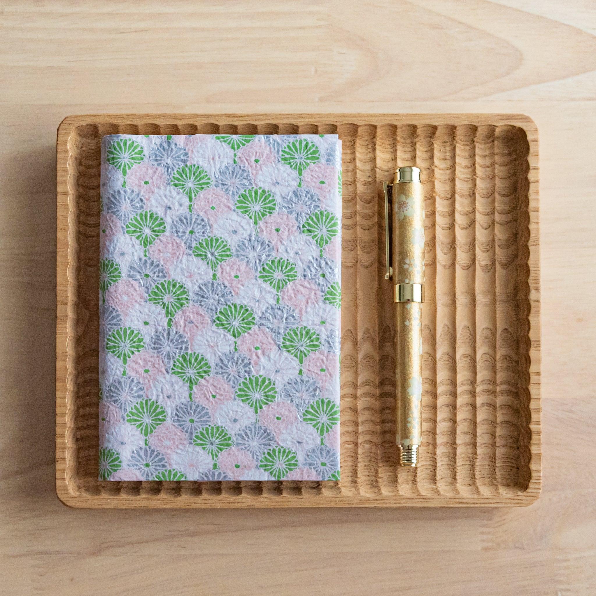 A notebook with a pink and green fan-like pattern and a matching gold pen, placed neatly on a wooden tray on a light wooden surface.