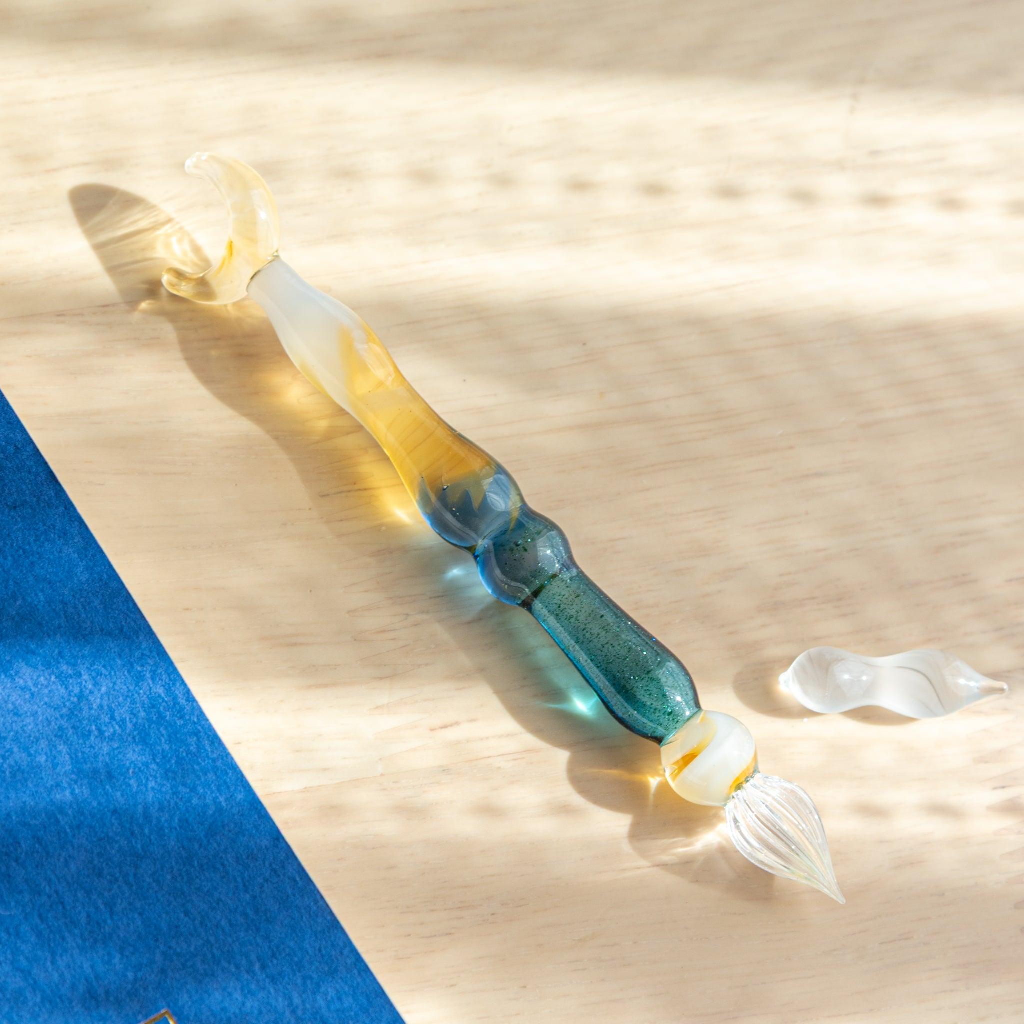 A blue and gold glass pen with a crescent moon top lies on a wooden surface. Its detachable nib is placed beside it in the sunlight.