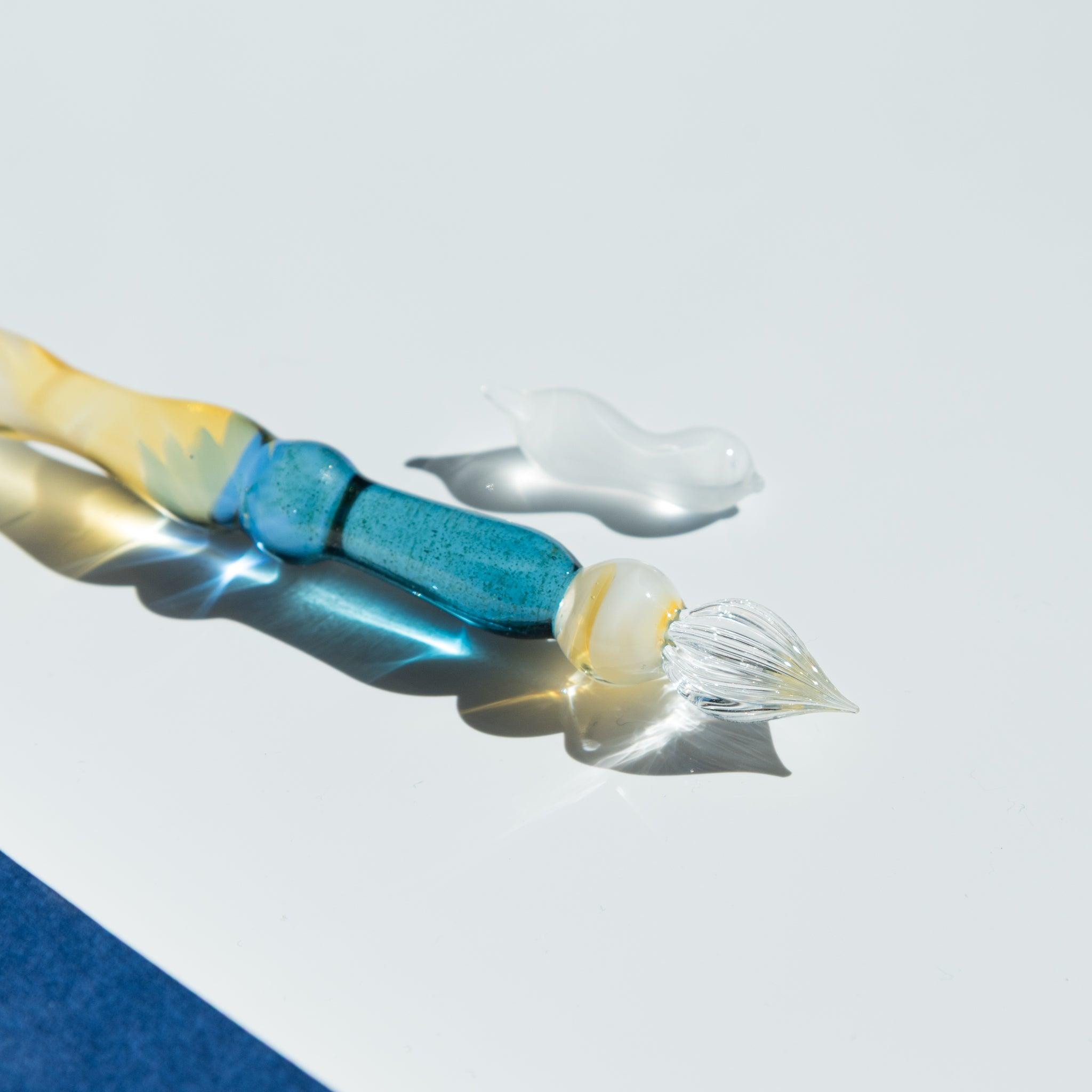 Close-up of the nib of a glass pen with a blue gradient body. The detachable nib and golden accents shimmer on a reflective white surface.