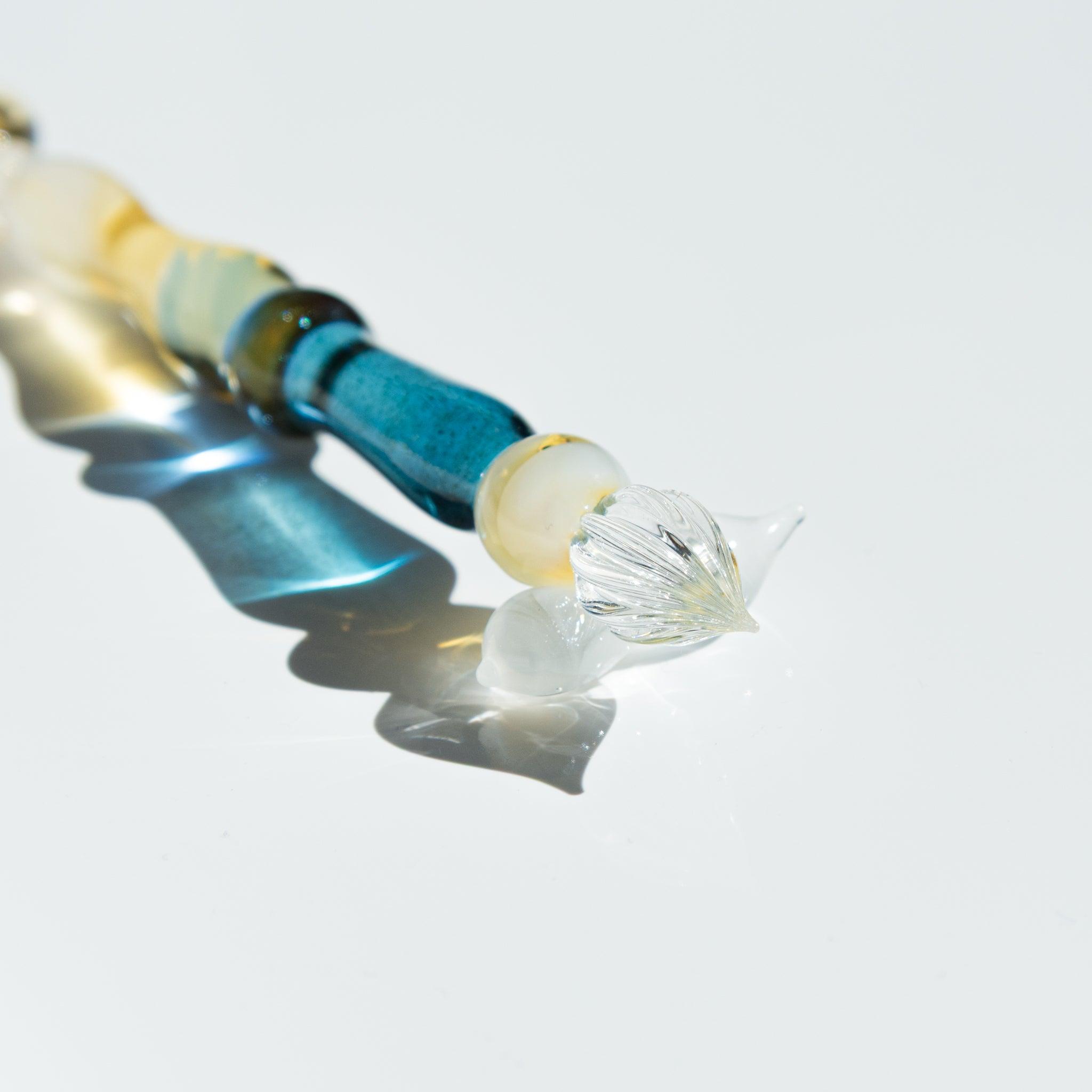 Extreme close-up of the nib of a glass pen with golden accents. The transparent glass catches the light on a reflective white surface.