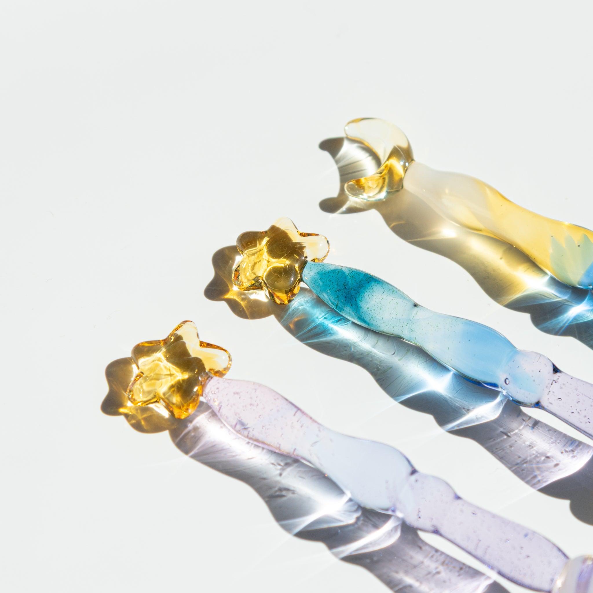 Close-up of the top parts of three glass pens with star and moon decorations. The transparent glass reflects light beautifully.