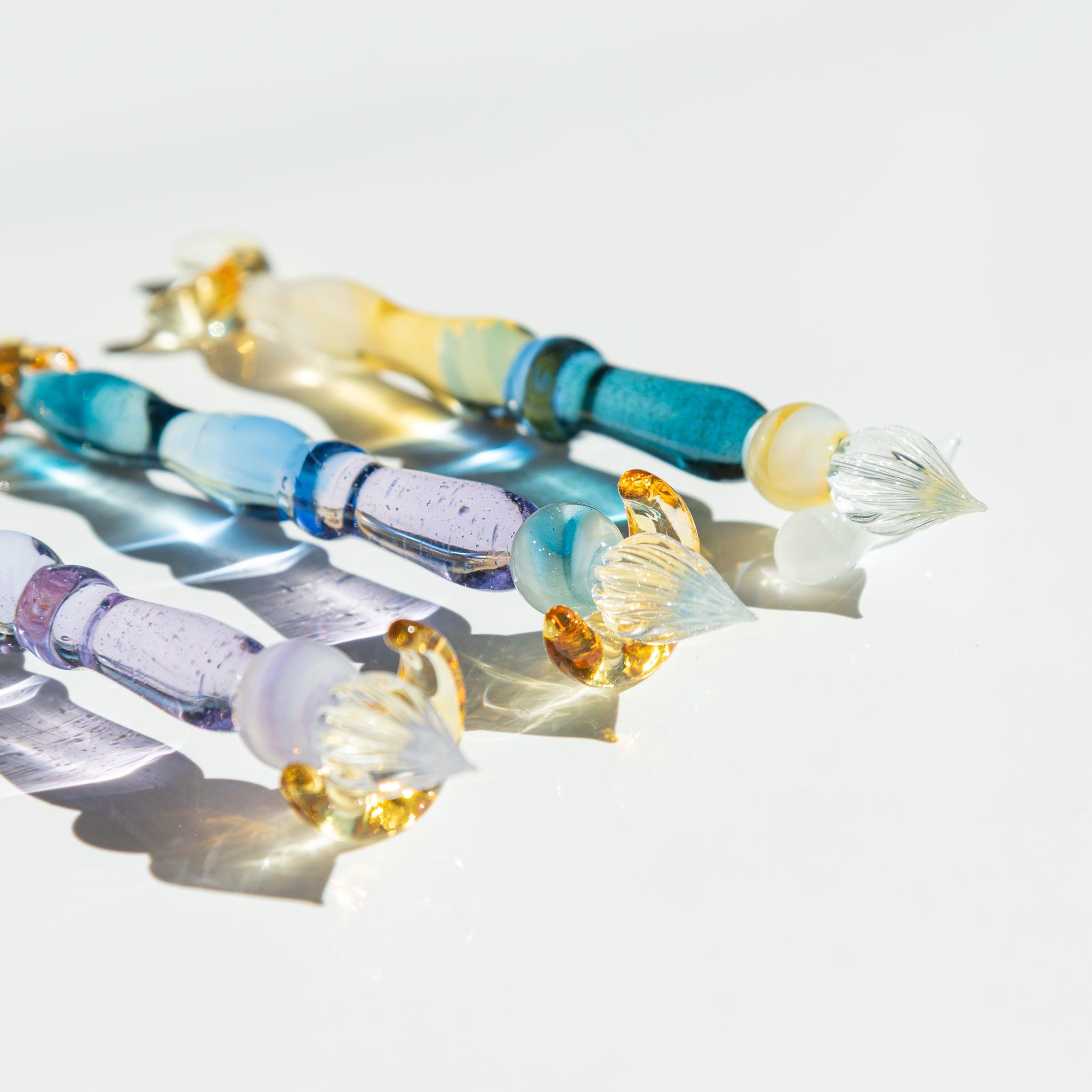 Close-up of the nibs of three glass pens with golden accents. The transparent blue and purple glass sparkles on a white surface.