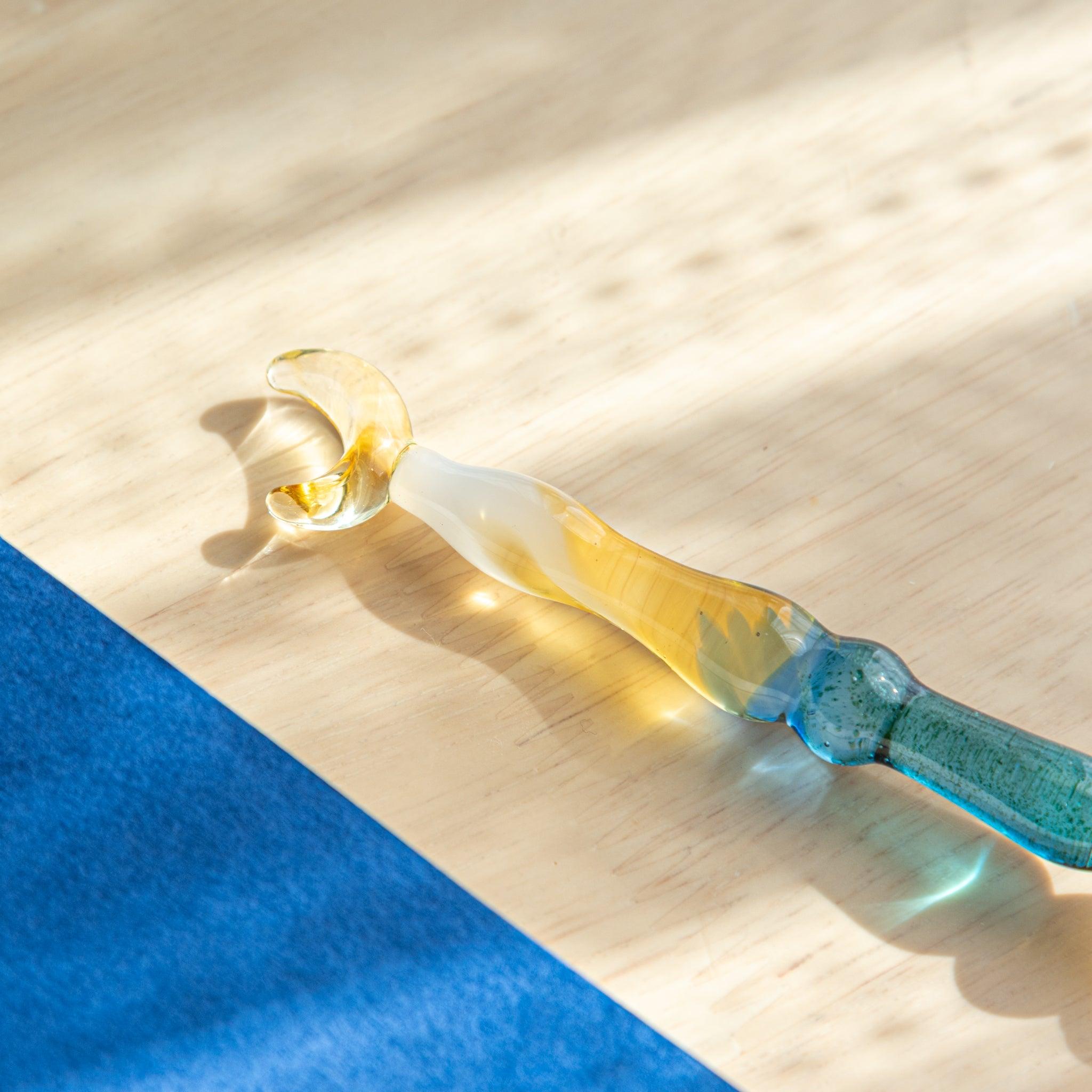 Close-up of a glass pen with a crescent moon top, resting on a wooden surface. The transparent blue and gold glass glows in the sunlight.