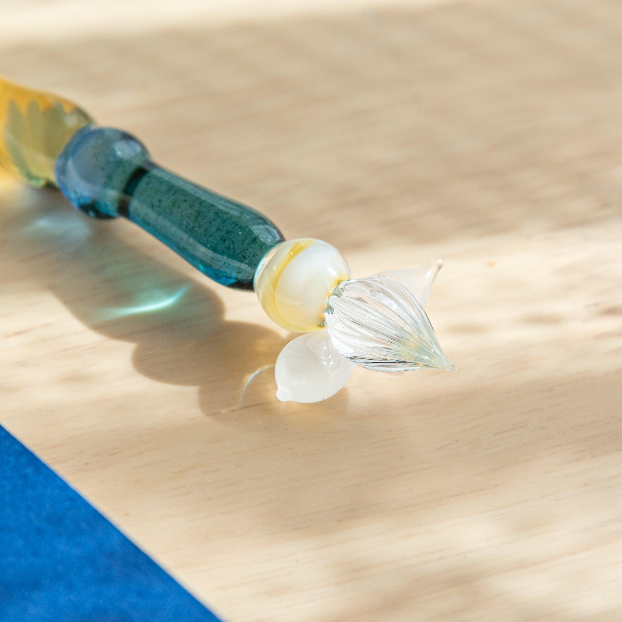 Close-up of the nib of a glass pen with golden accents. The transparent glass reflects sunlight on a wooden surface with a blue notebook nearby.