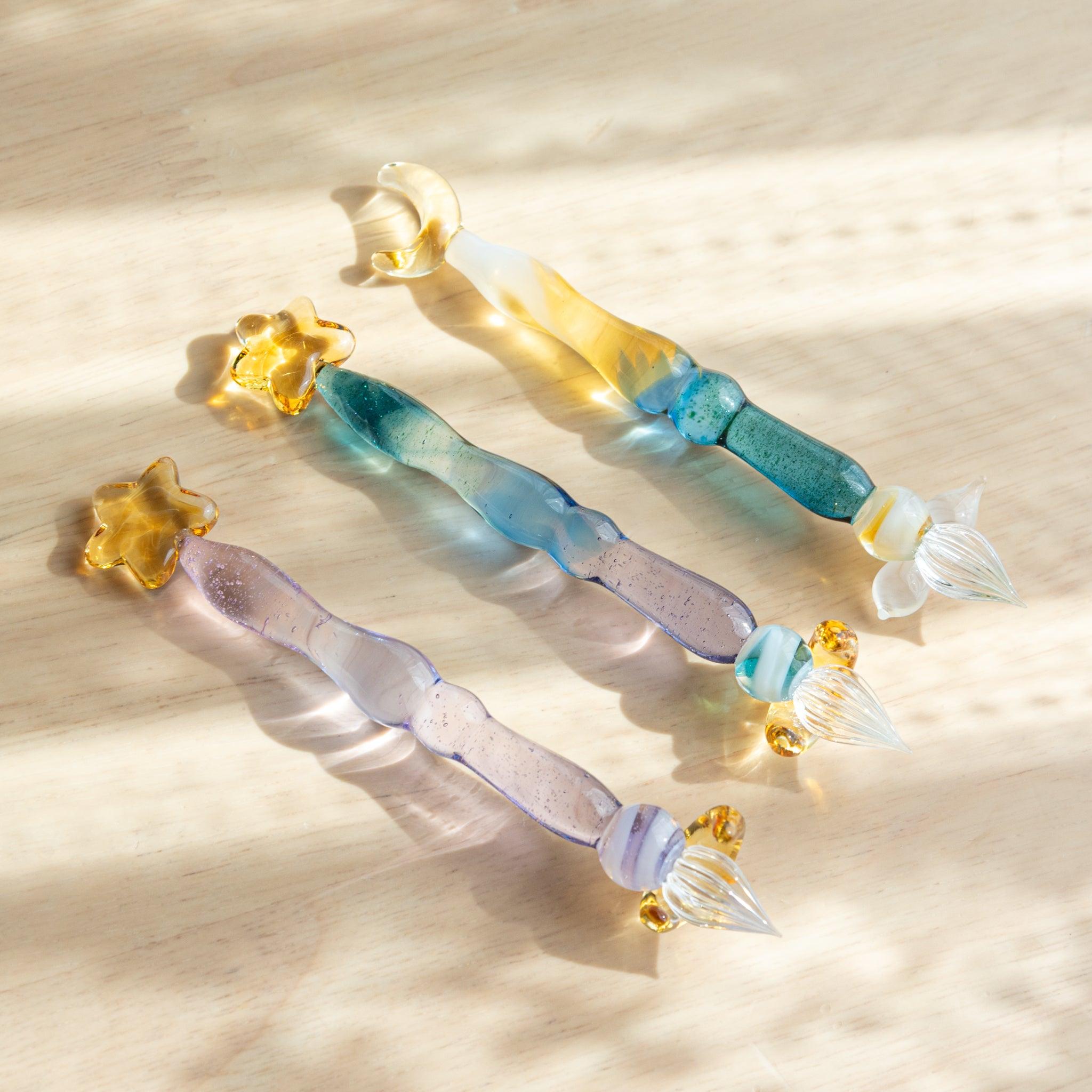 Three glass pens with star and moon decorations placed on a wooden surface. The sunlight highlights their transparent blue and purple hues.