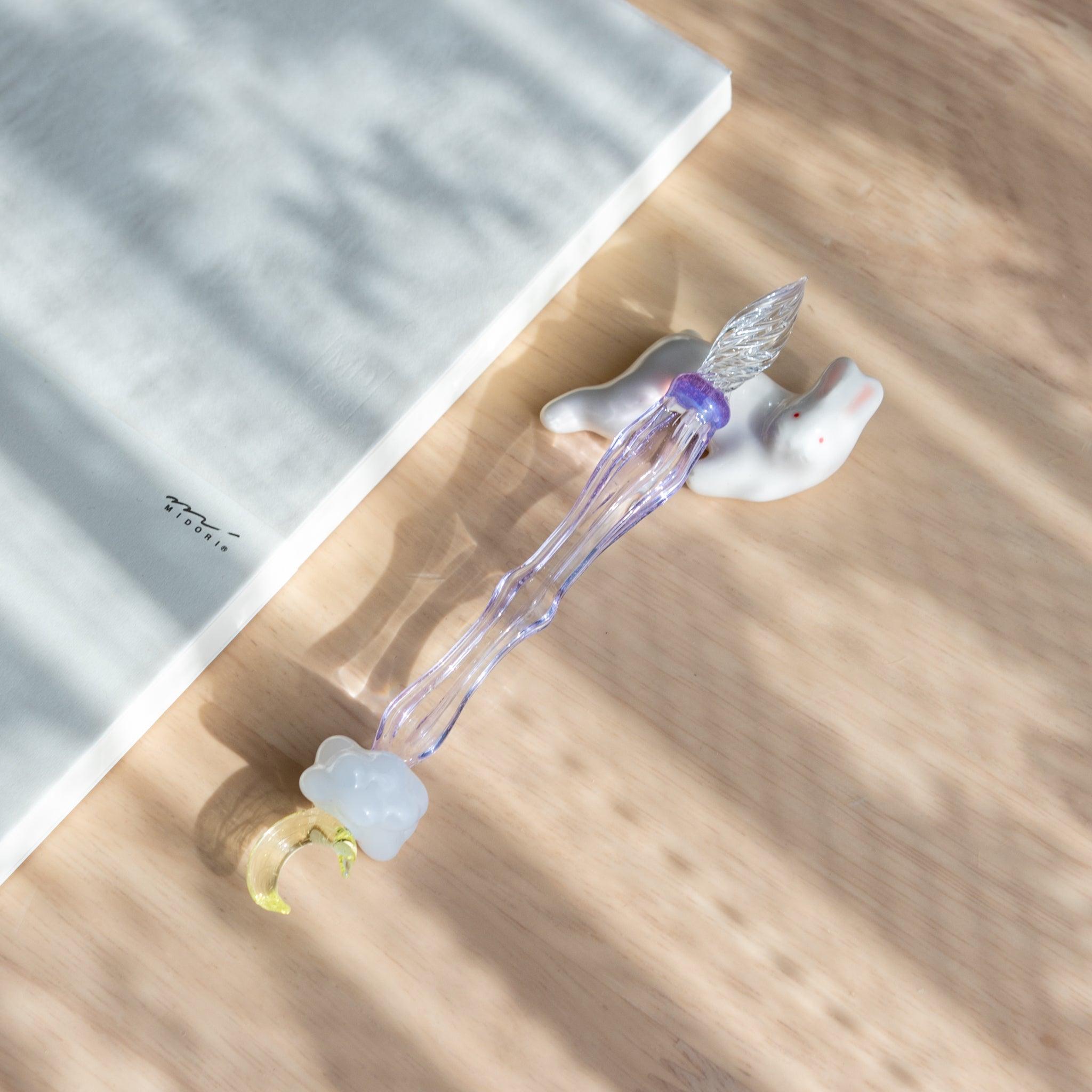 A handmade ceramic pen rest shaped as a white rabbit, holding a purple glass dip pen with a moon motif. Part of traditional Japanese stationery.