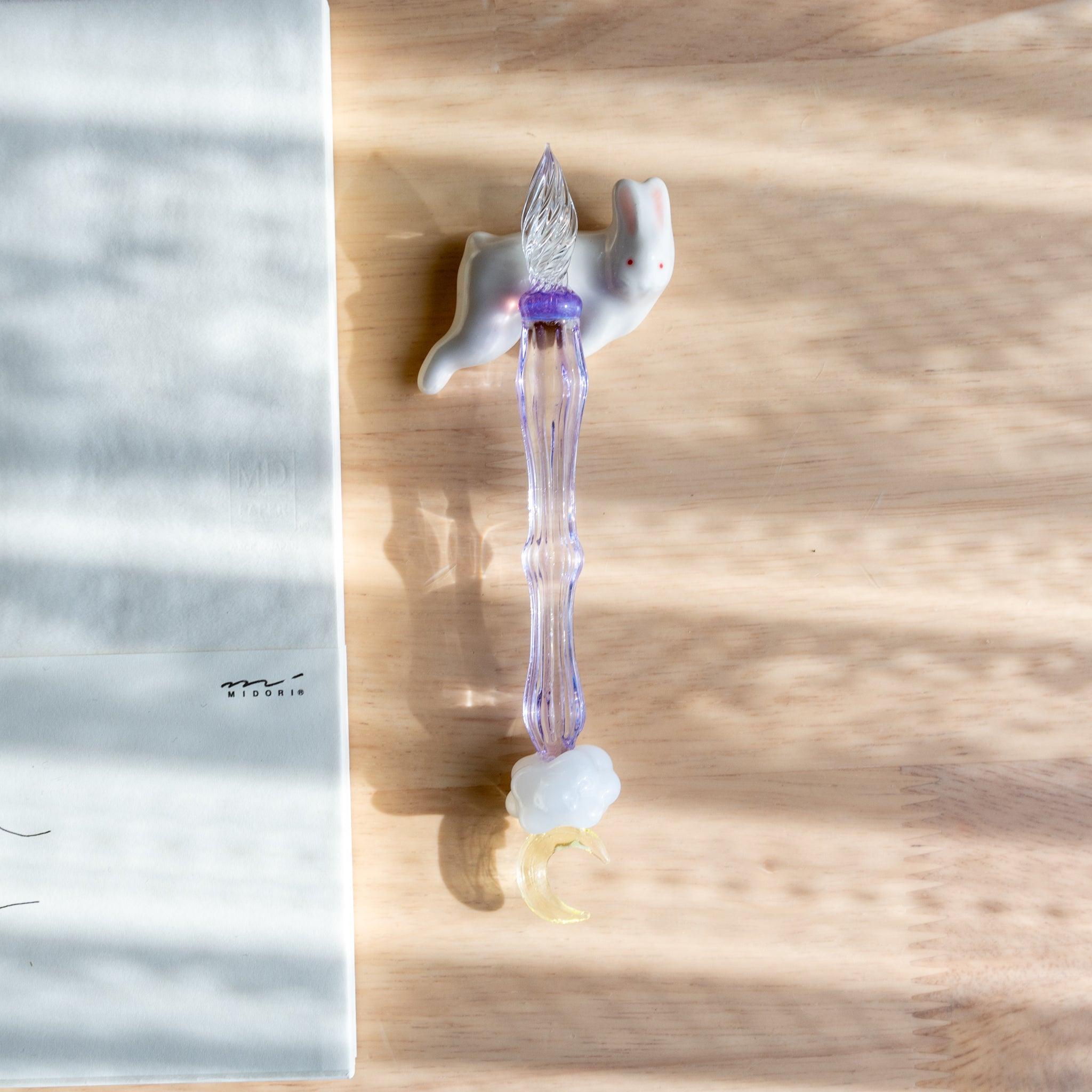 A handmade ceramic pen rest shaped as a white rabbit, holding a purple glass dip pen with a moon motif. Part of traditional Japanese stationery.