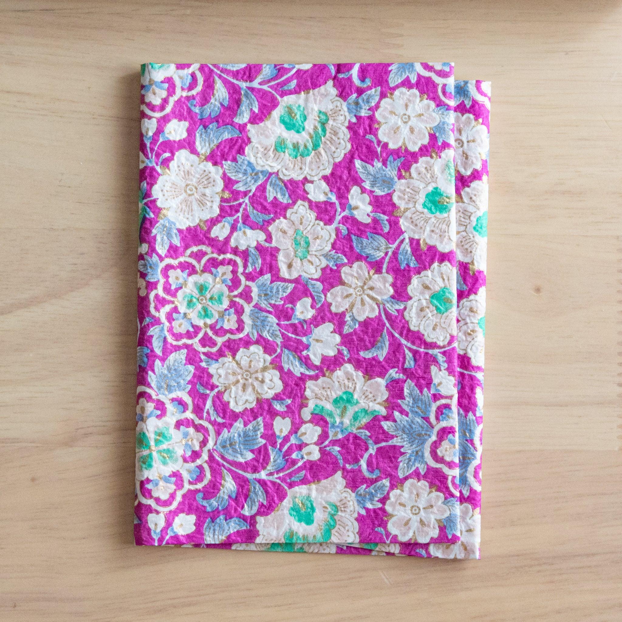 A notebook with a vibrant pink floral pattern featuring intricate white and green flowers, placed on a light wooden surface.