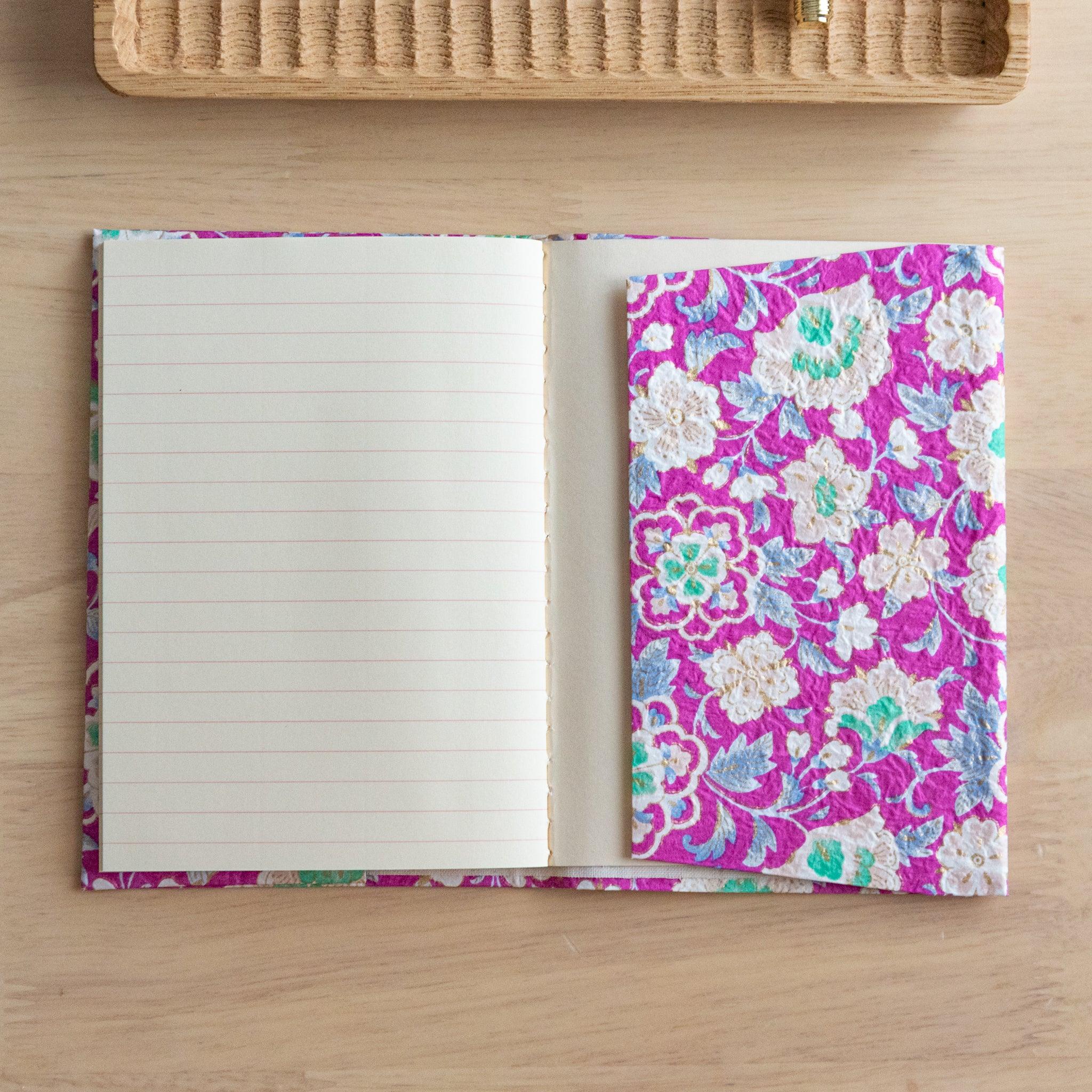 An open notebook with a vibrant pink floral cover, showing a lined page and an inner pocket, placed on a wooden surface.