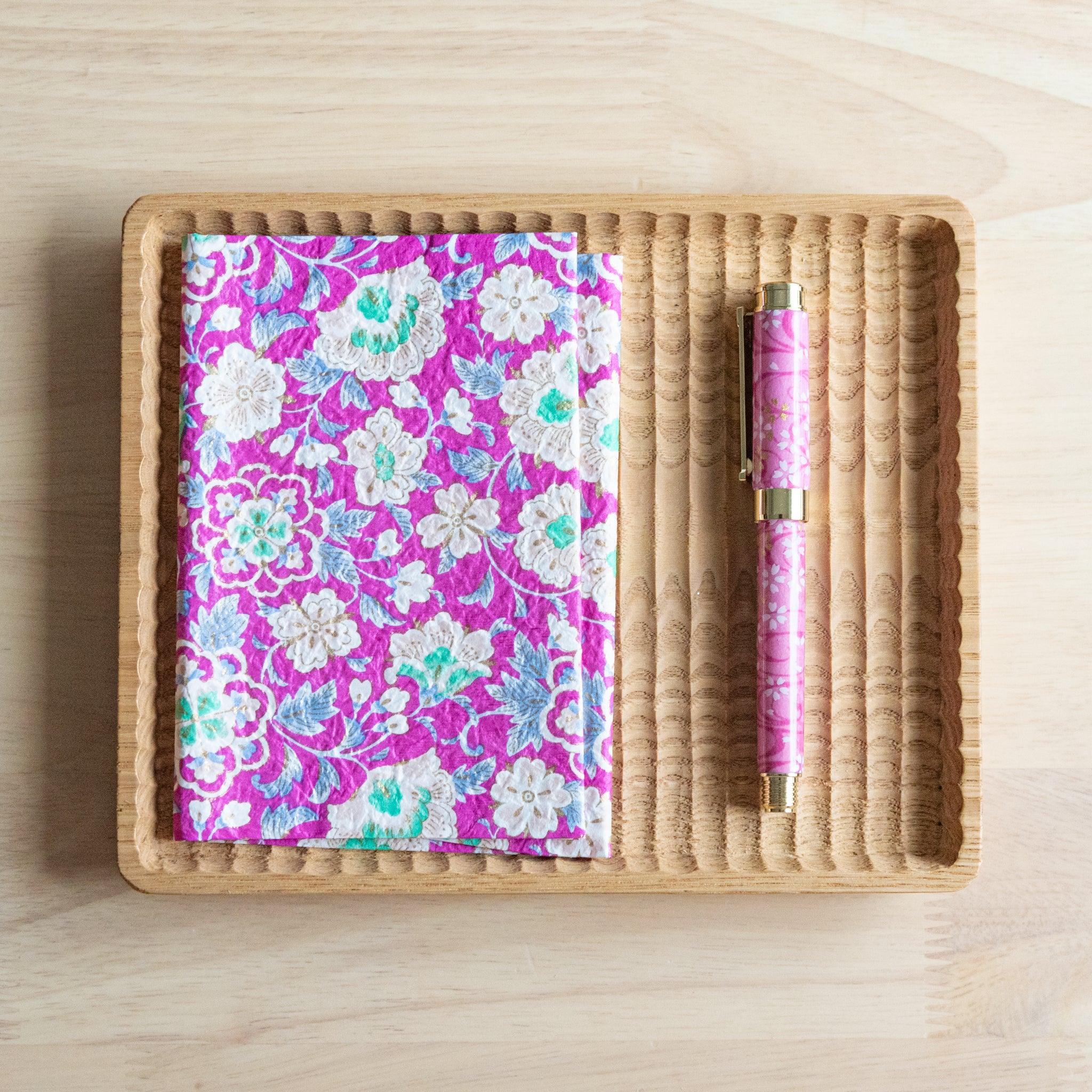 A notebook with a vibrant pink floral pattern and a matching pink pen, neatly arranged on a wooden tray on a light wooden surface.