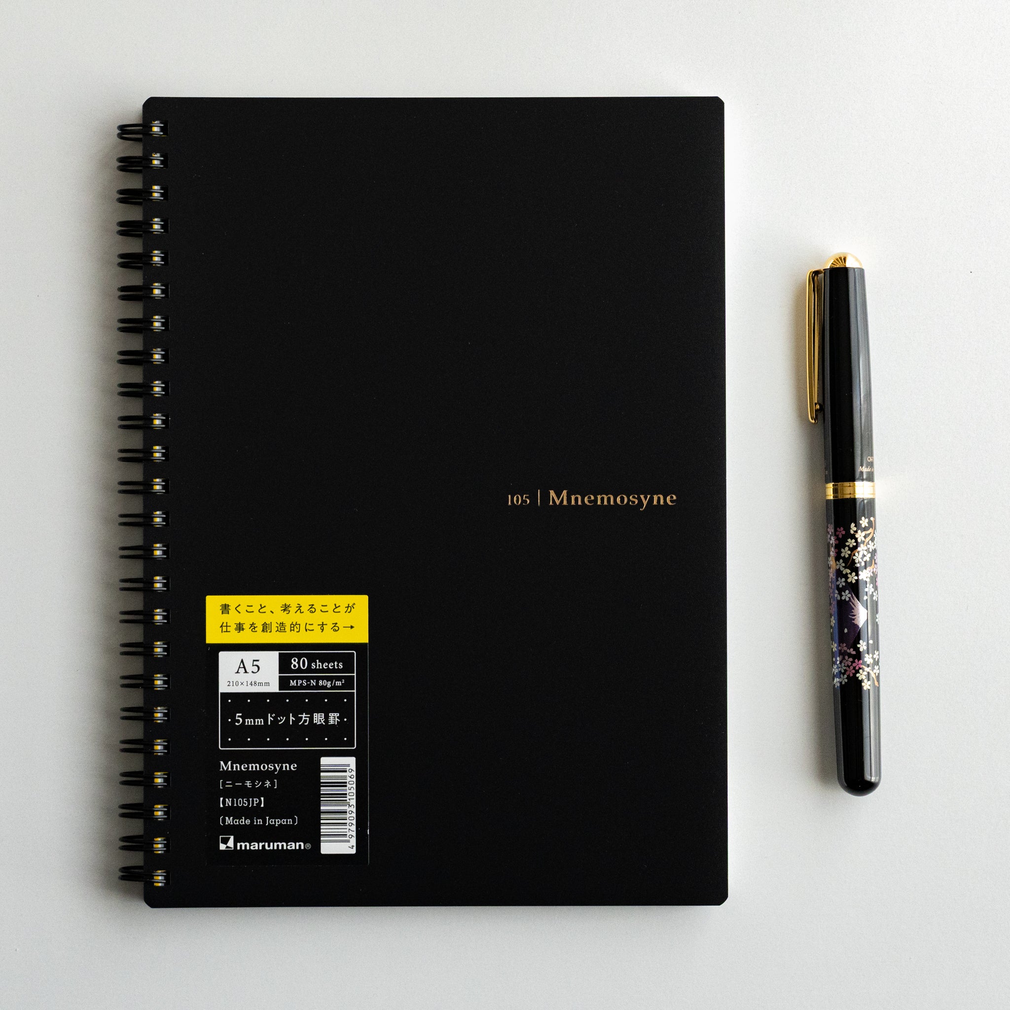 Maruman Mnemosyne spiral-bound dot grid notebook with black cover and yellow label, displayed next to a pen for scale.