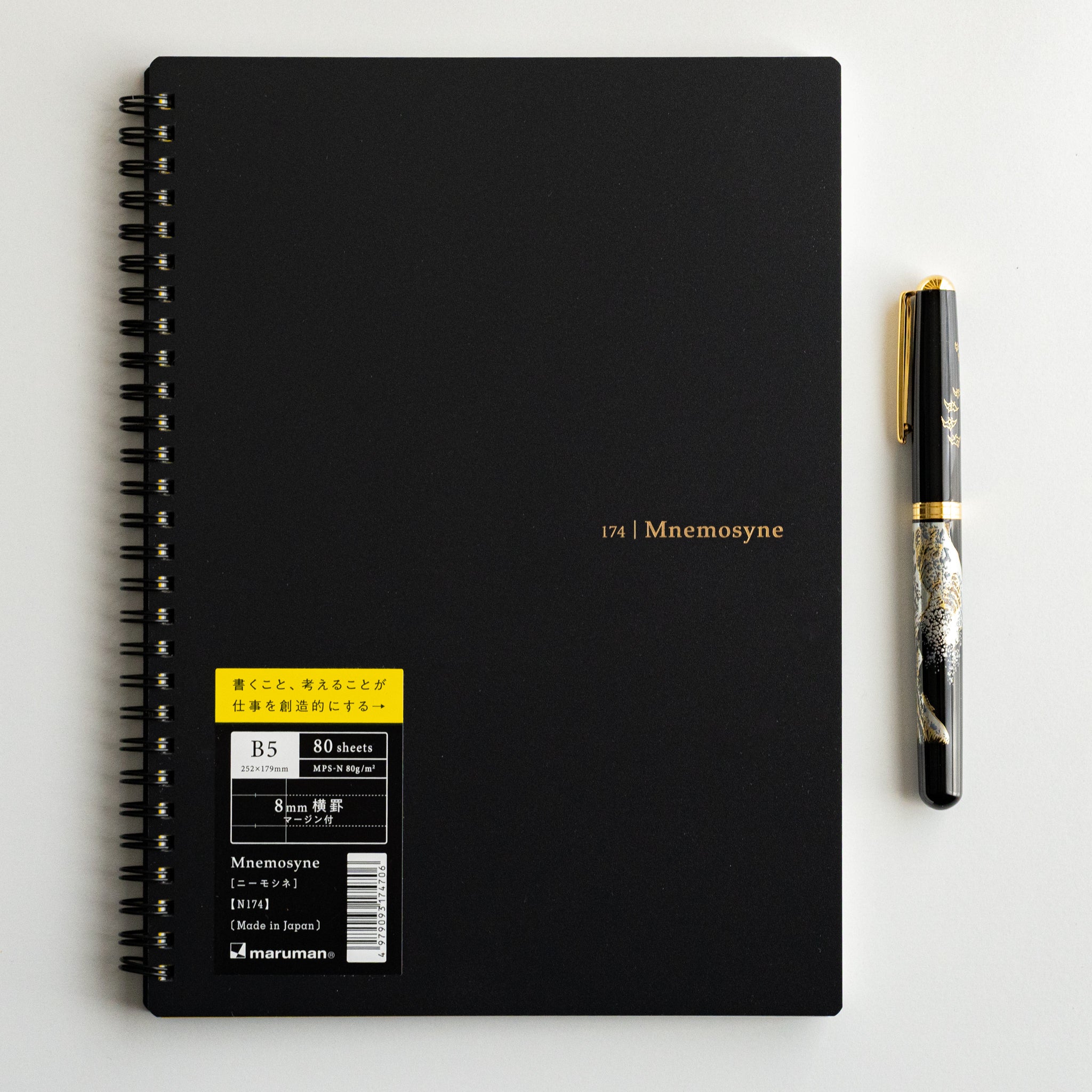 Maruman Mnemosyne lined notebook with black cover and yellow product label, displayed beside a sleek pen for a refined workspace aesthetic.