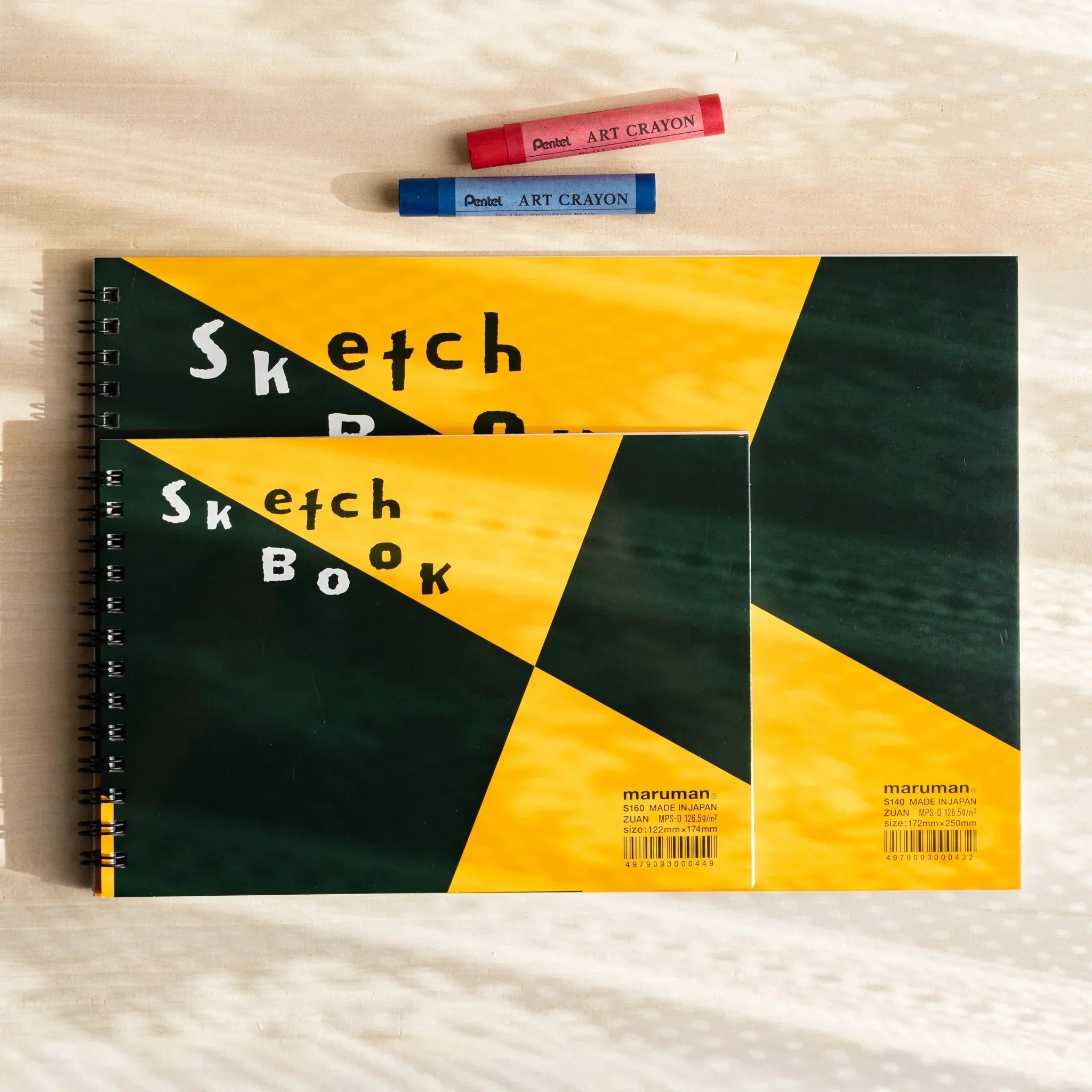 Two Maruman Japanese sketchbooks with yellow and green ring-bound covers on a desk, with two crayons for arts and crafts.