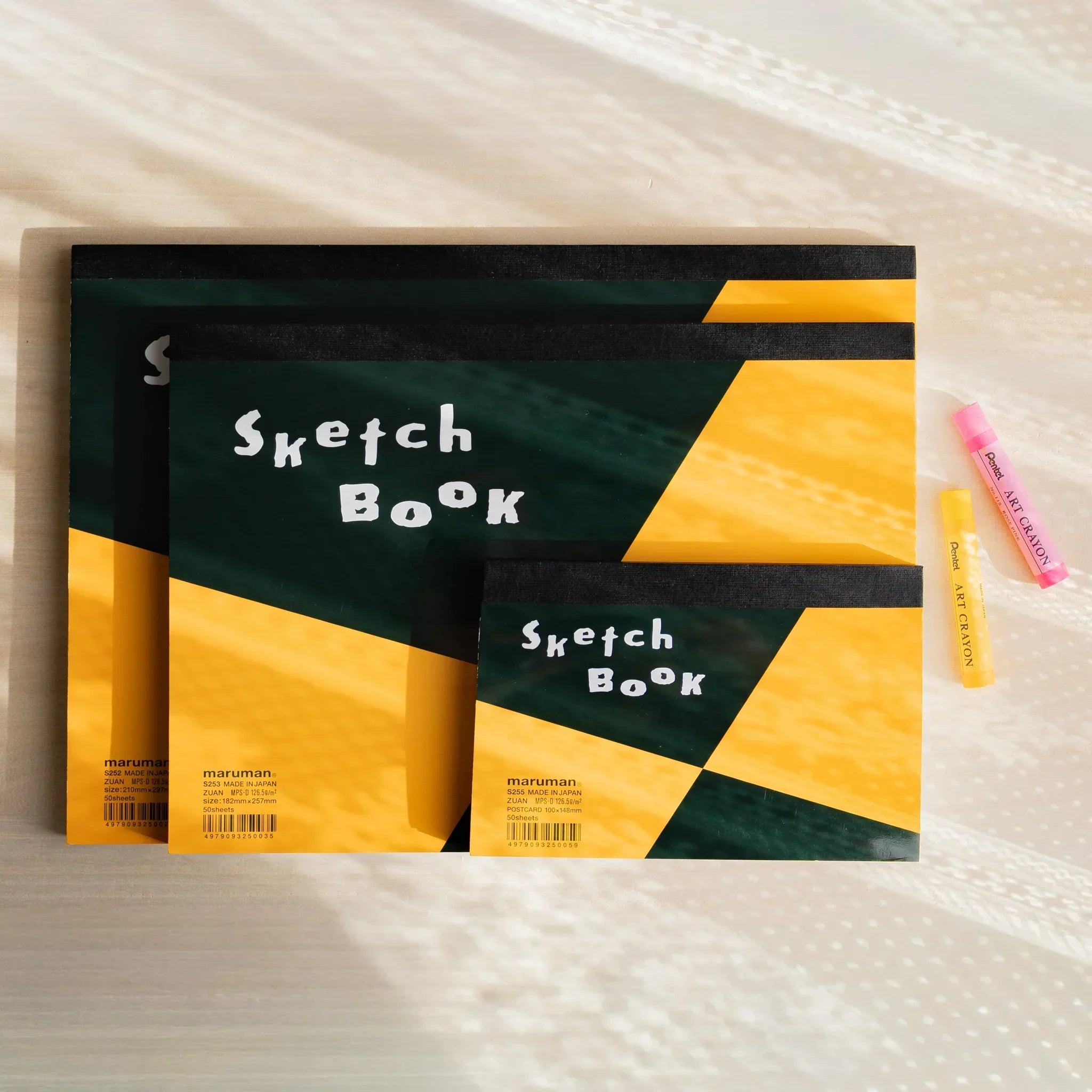 Maruman Japanese sketchbook pads with spiral binding, orange and black covers, shown with pastel crayons for arts and crafts.