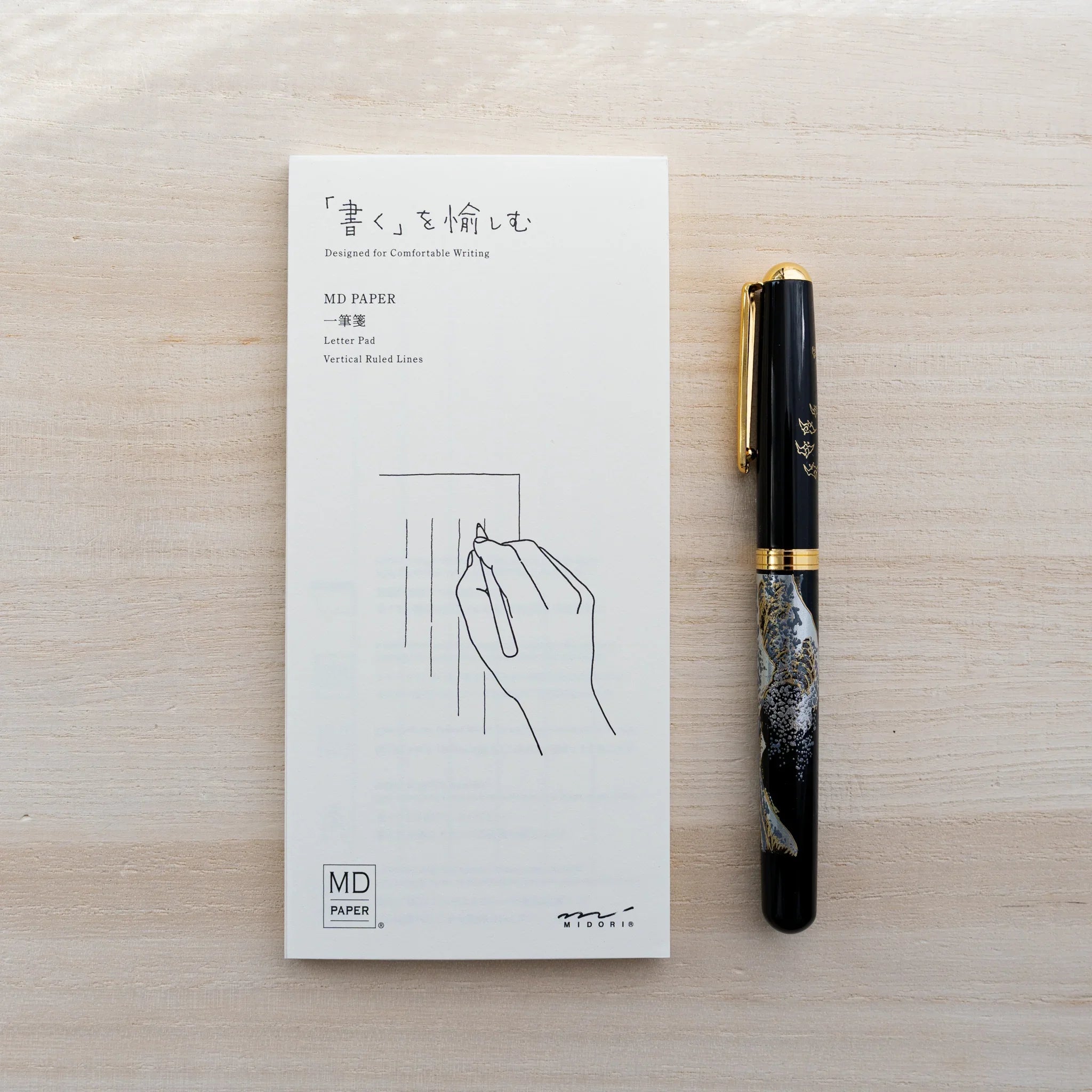 White MD Paper letter pad with hand illustration beside a black fountain pen with gold trim on wood