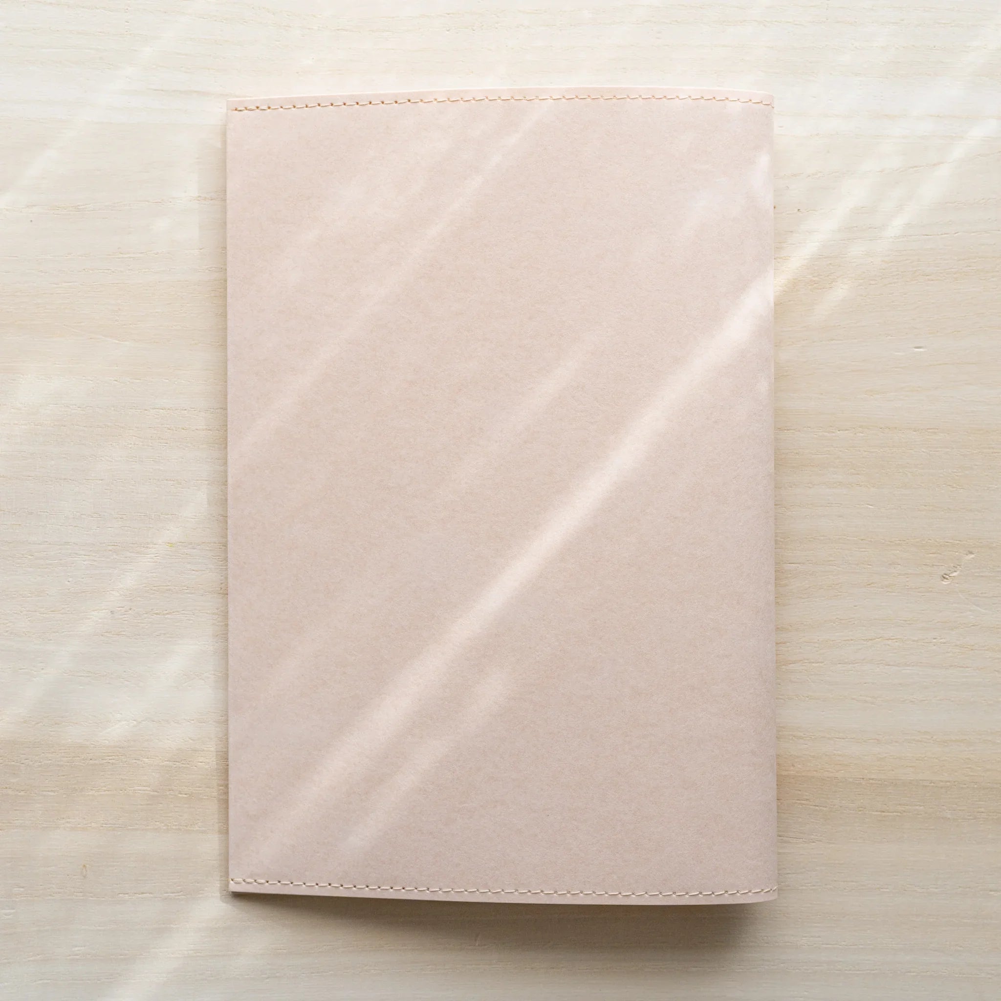 Midori MD Notebook Paper Cover front view from above, plain pale pink paper with stitched edges, no belly band on wood