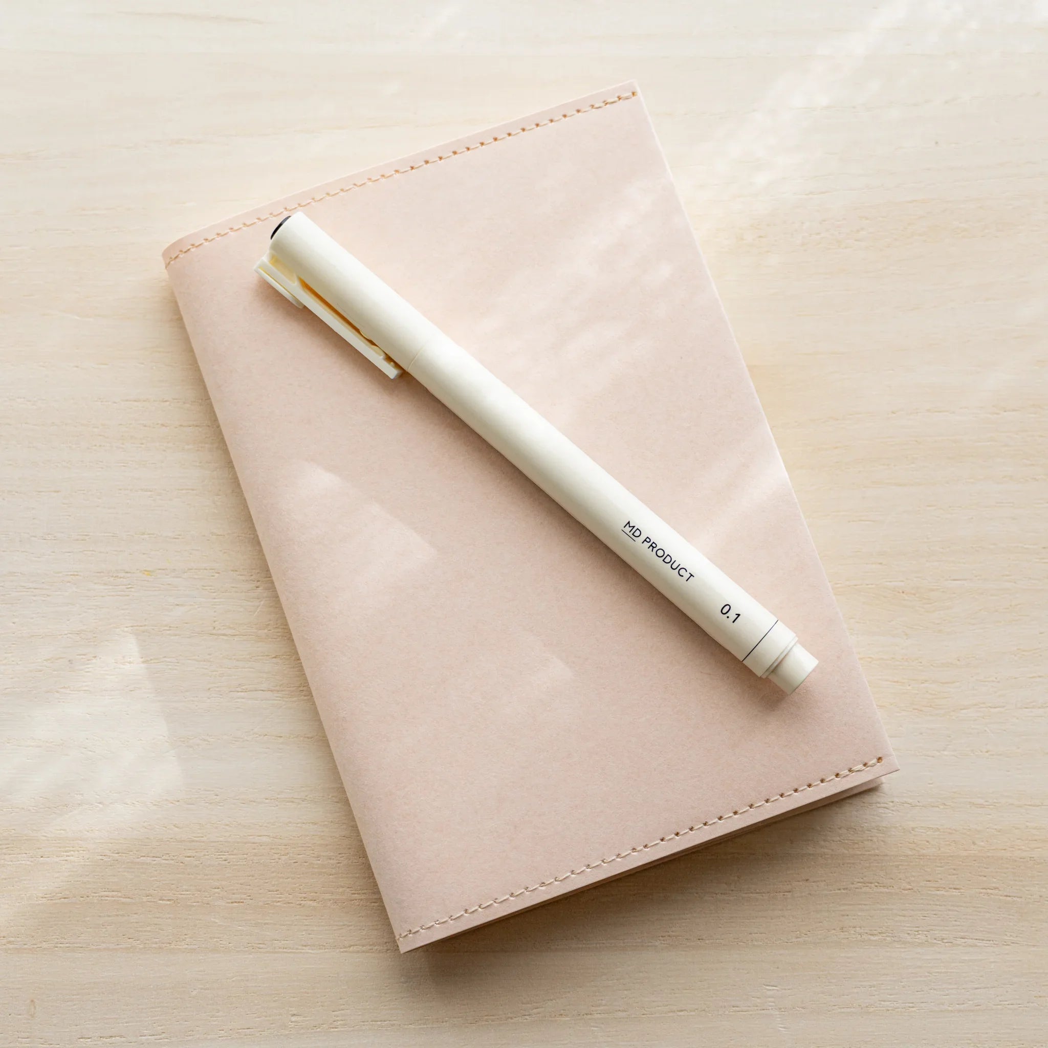 Pale pink MD Notebook Paper Cover with a pen resting on top, closed view on wood