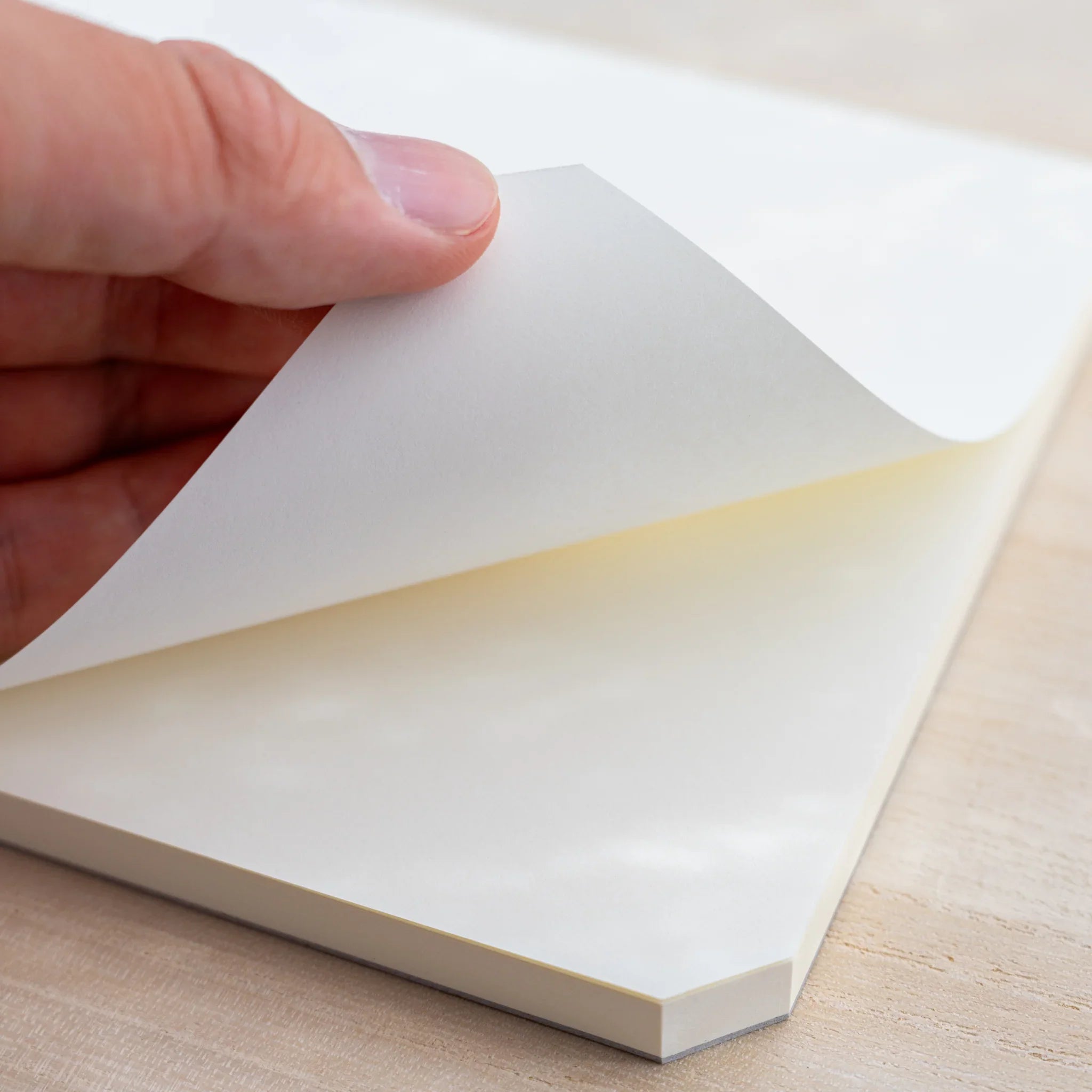 A hand lifting the blank cream top sheet of a Midori MD Paper Pad A5, cut corner and stacked sheets visible