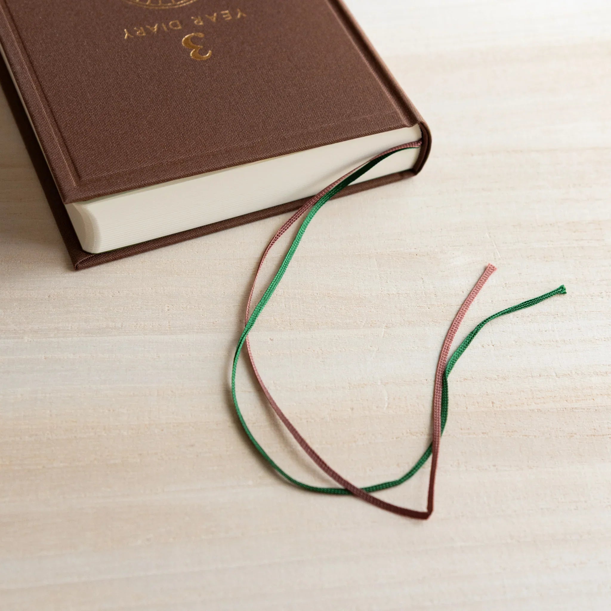 Close-up of brown Midori Japanese Journal with dual green and brown bookmarks on a light wooden desk.