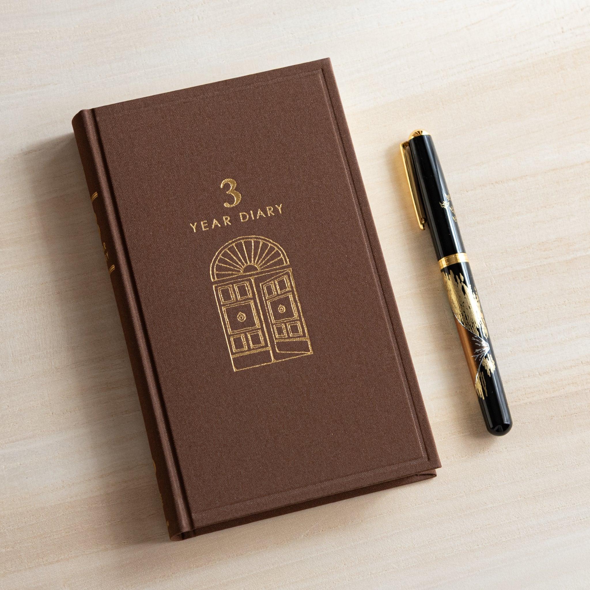 Brown Midori Japanese journal 3 Year Diary Gate Design with gold embossed door motif beside black and gold fountain pen.