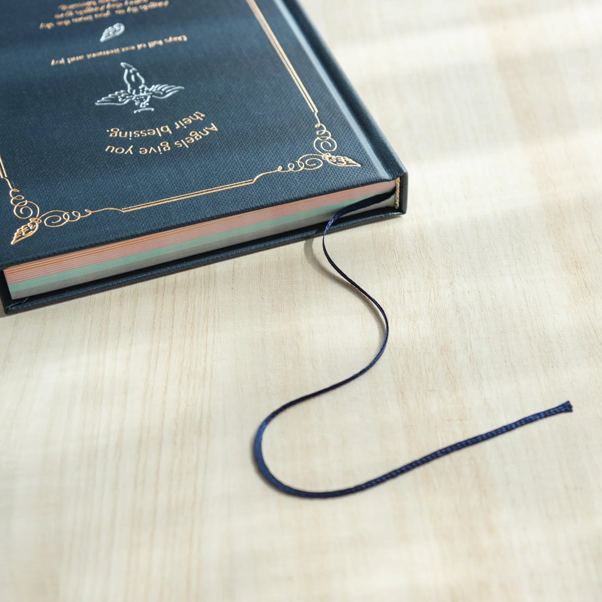 Midori Angel Motif Journal closed on wooden desk showing gold foil cover design, pastel page edges, and navy ribbon bookmark
