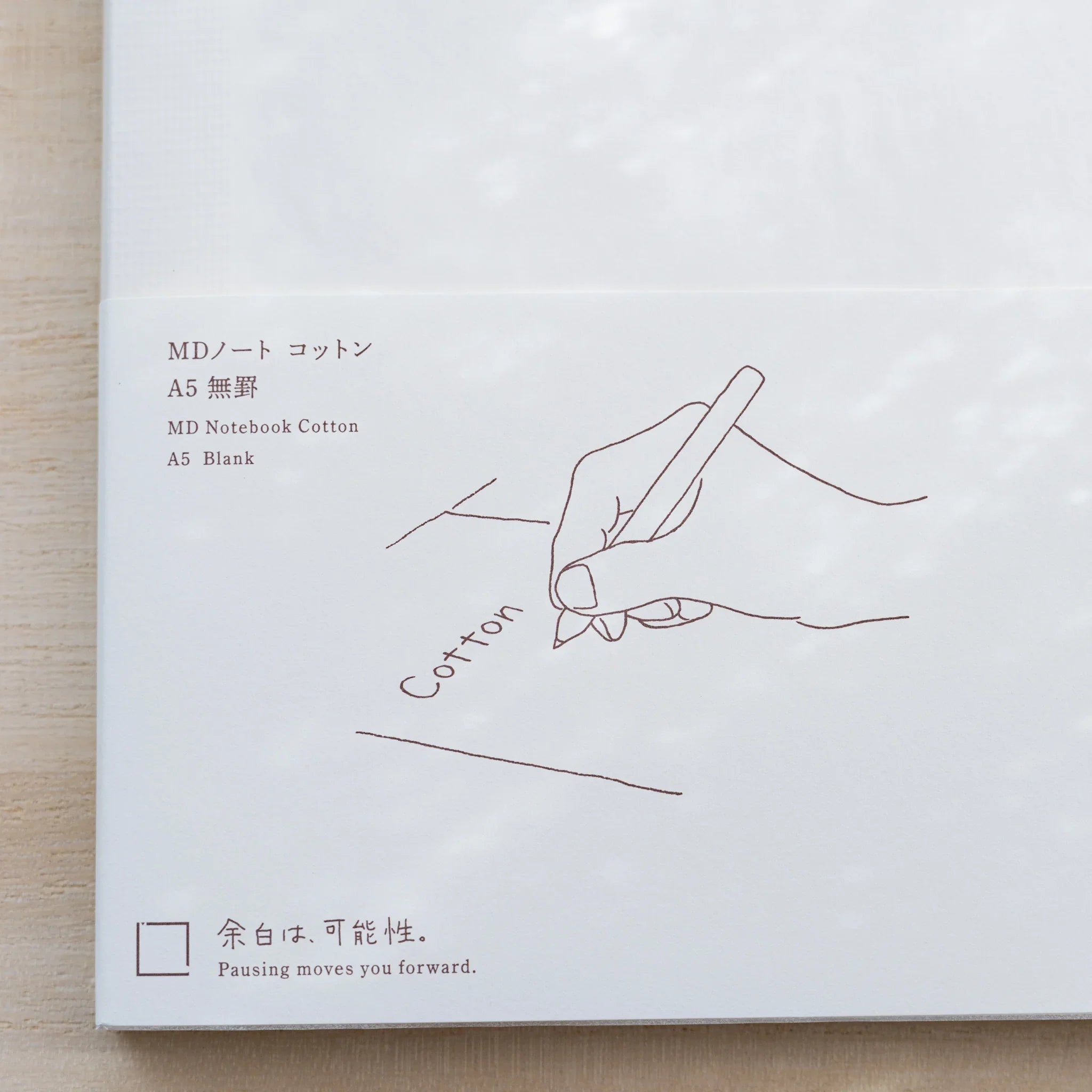 Close-up of Midori MD Cotton Notebook A5 belly band in brown, line drawing of a hand writing the word Cotton