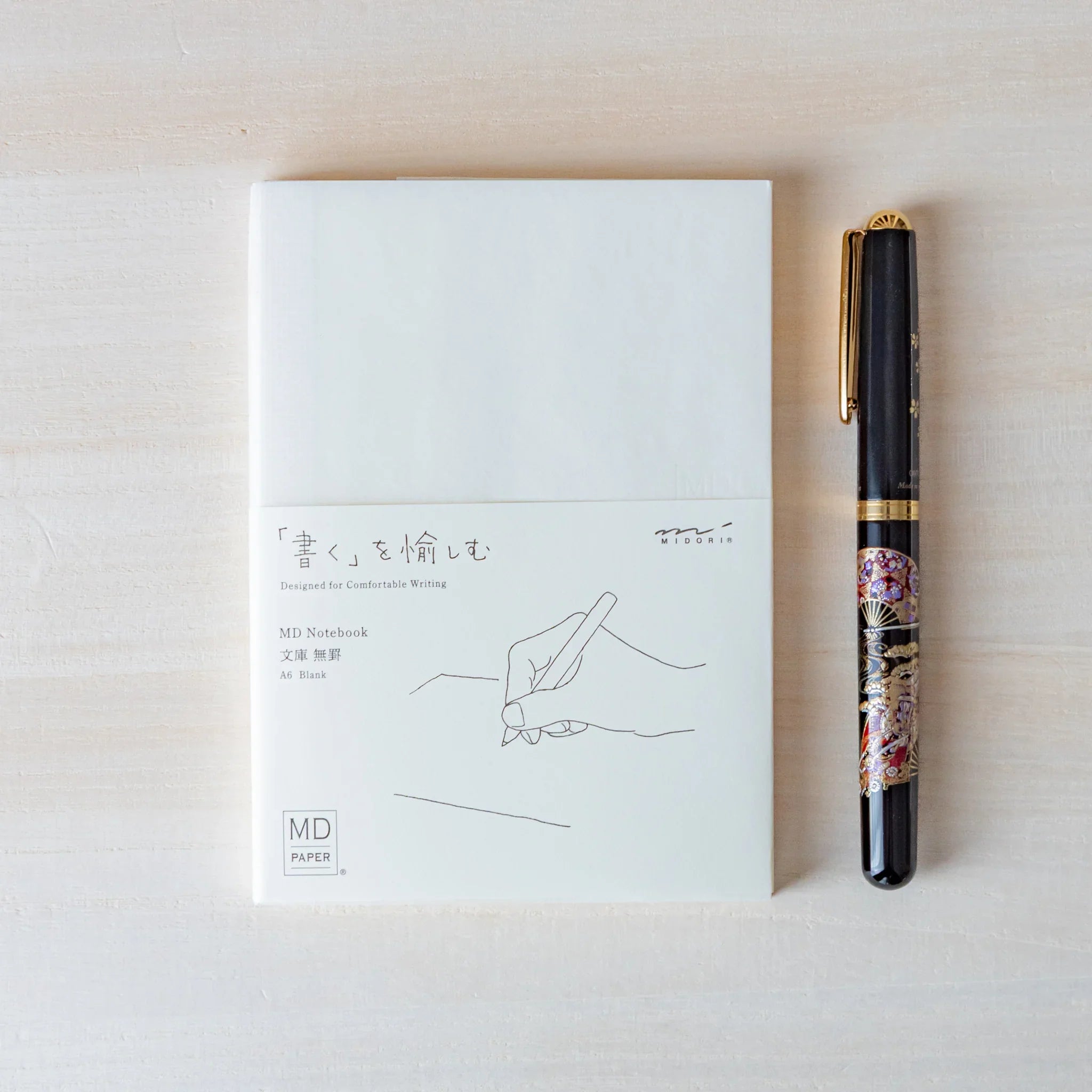 Japanese Notebook Midori MD A6 notebook with white cover placed beside a fountain pen on a light wooden surface.