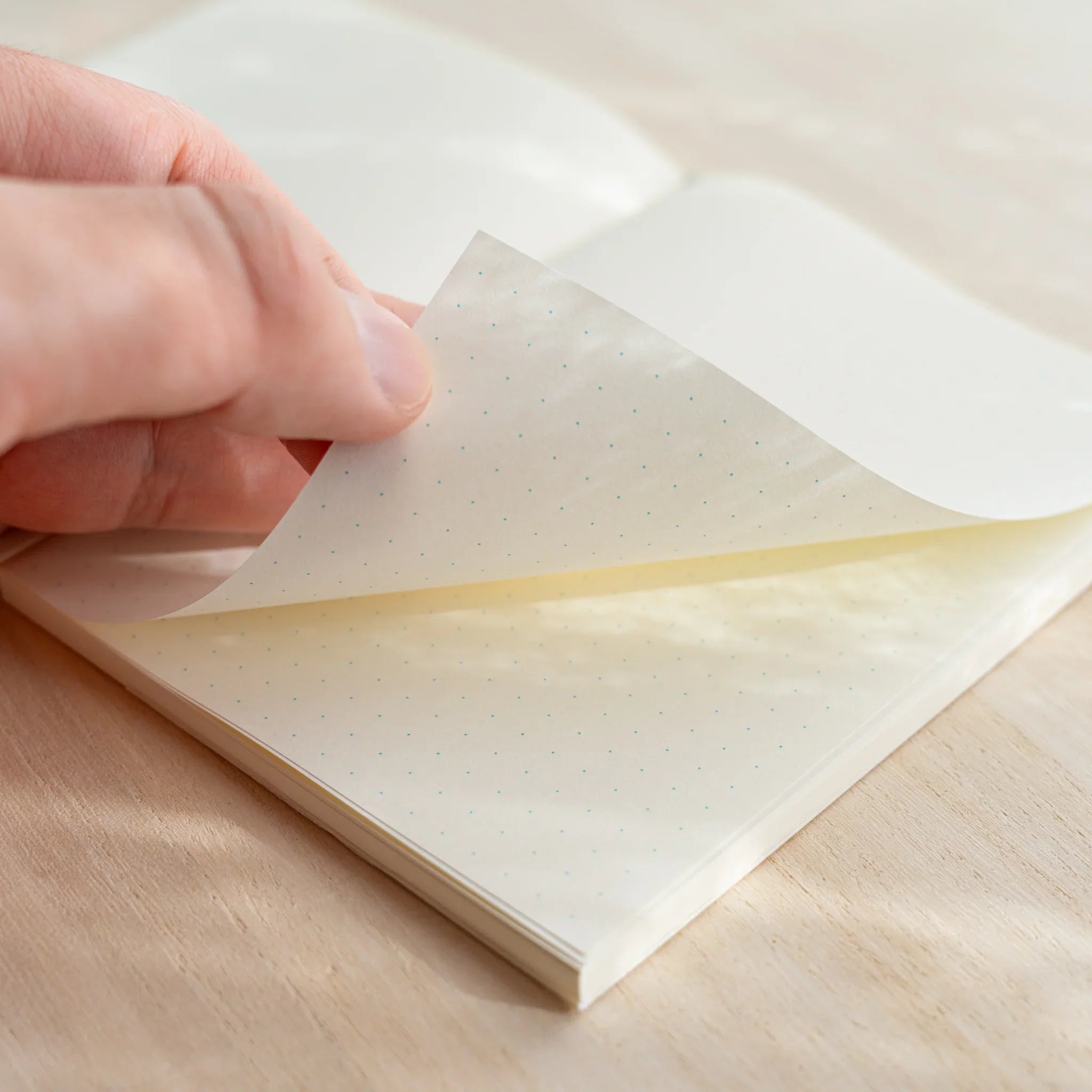 Hand lifting glassine cover of Midori MD Notebook A6, revealing ivory dot grid pages underneath, showing paper thinness