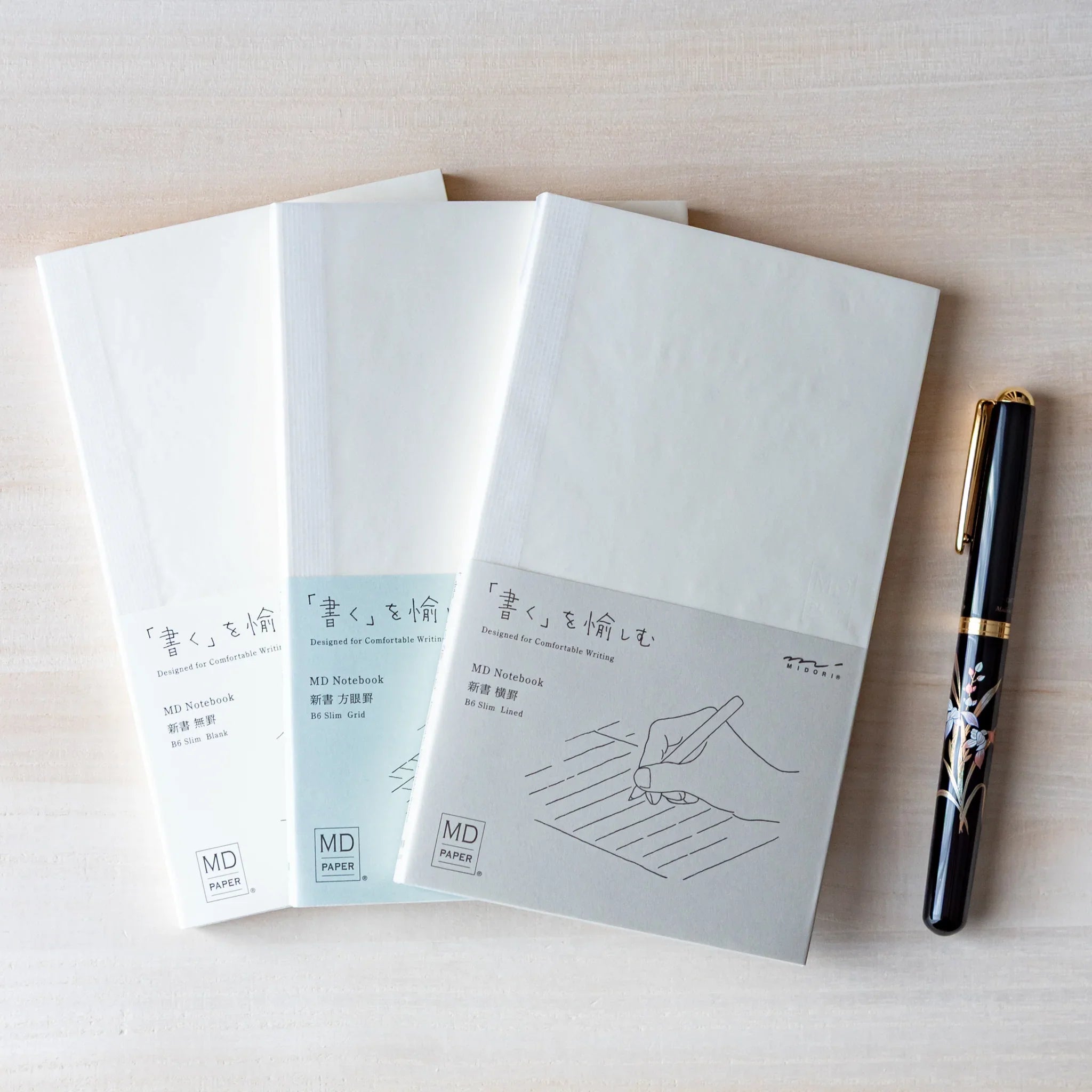 Japanese Notebook set: three Midori MD B6 notebooks with minimalist white covers and a black pen on a light wooden surface.