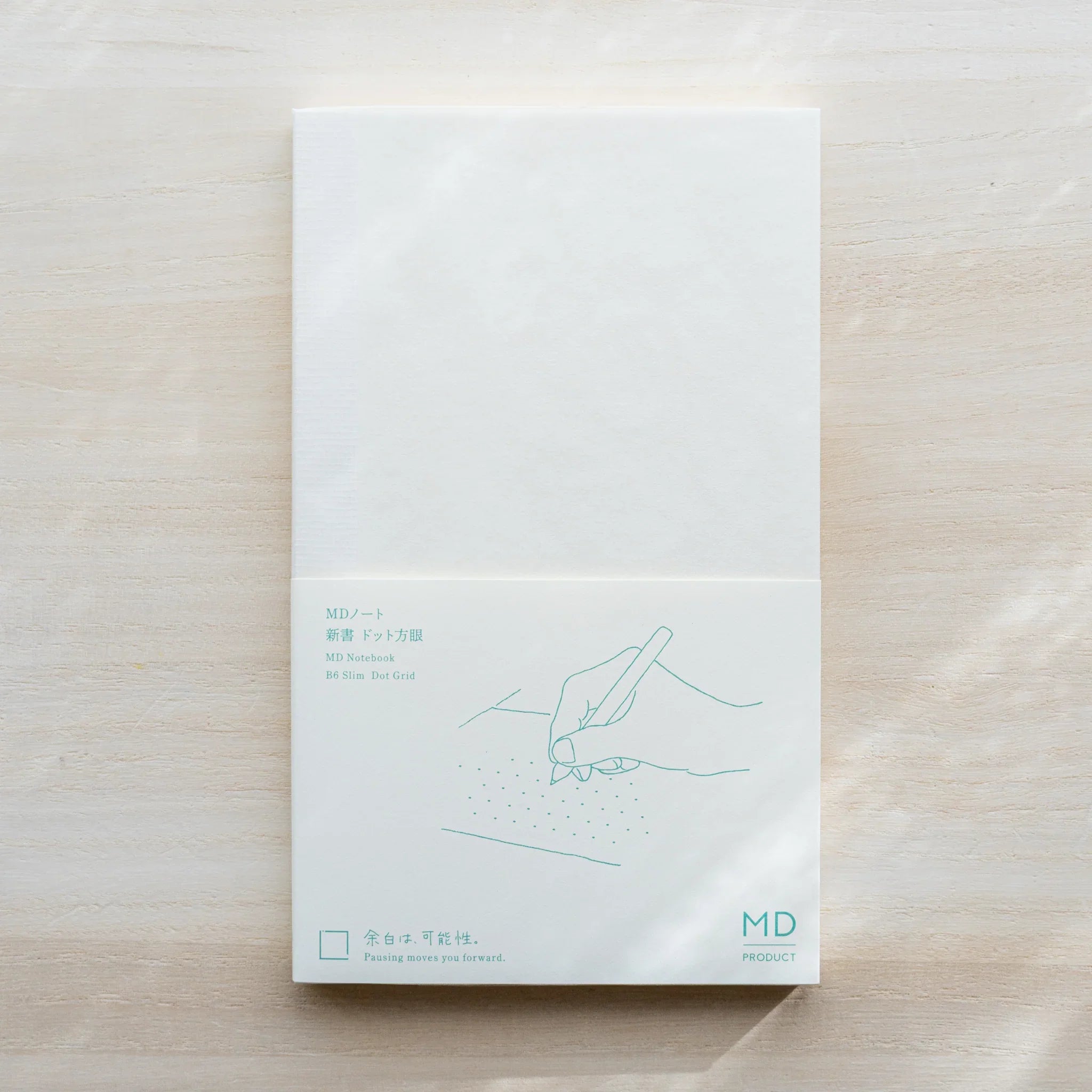 Midori MD Notebook B6 Slim Dot Grid on light wood surface, white cover with teal hand illustration and MD PRODUCT logo