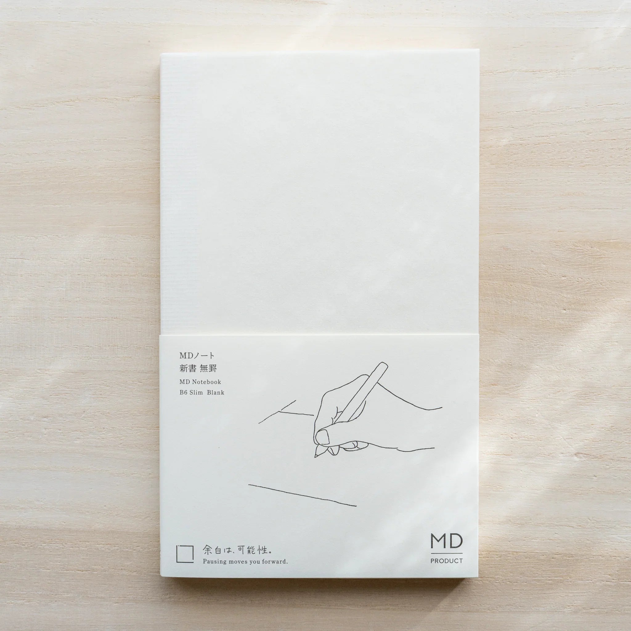 Midori MD Notebook B6 Slim Blank on light wood, white cover with hand and pen illustration and MD PRODUCT logo