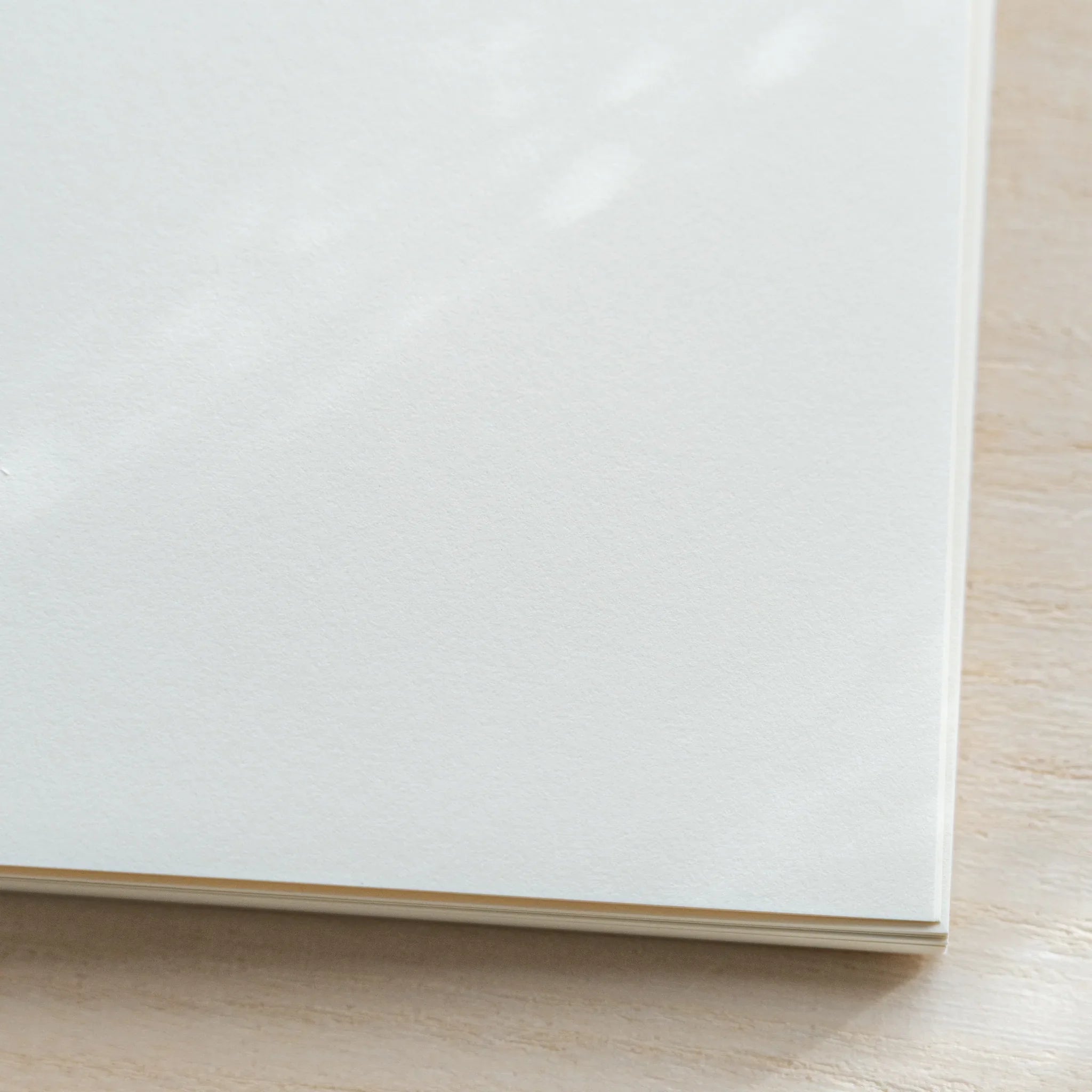 Close-up of Midori MD Notebook B6 Slim corner, plain white cover, page edges visible at bottom, on wood surface