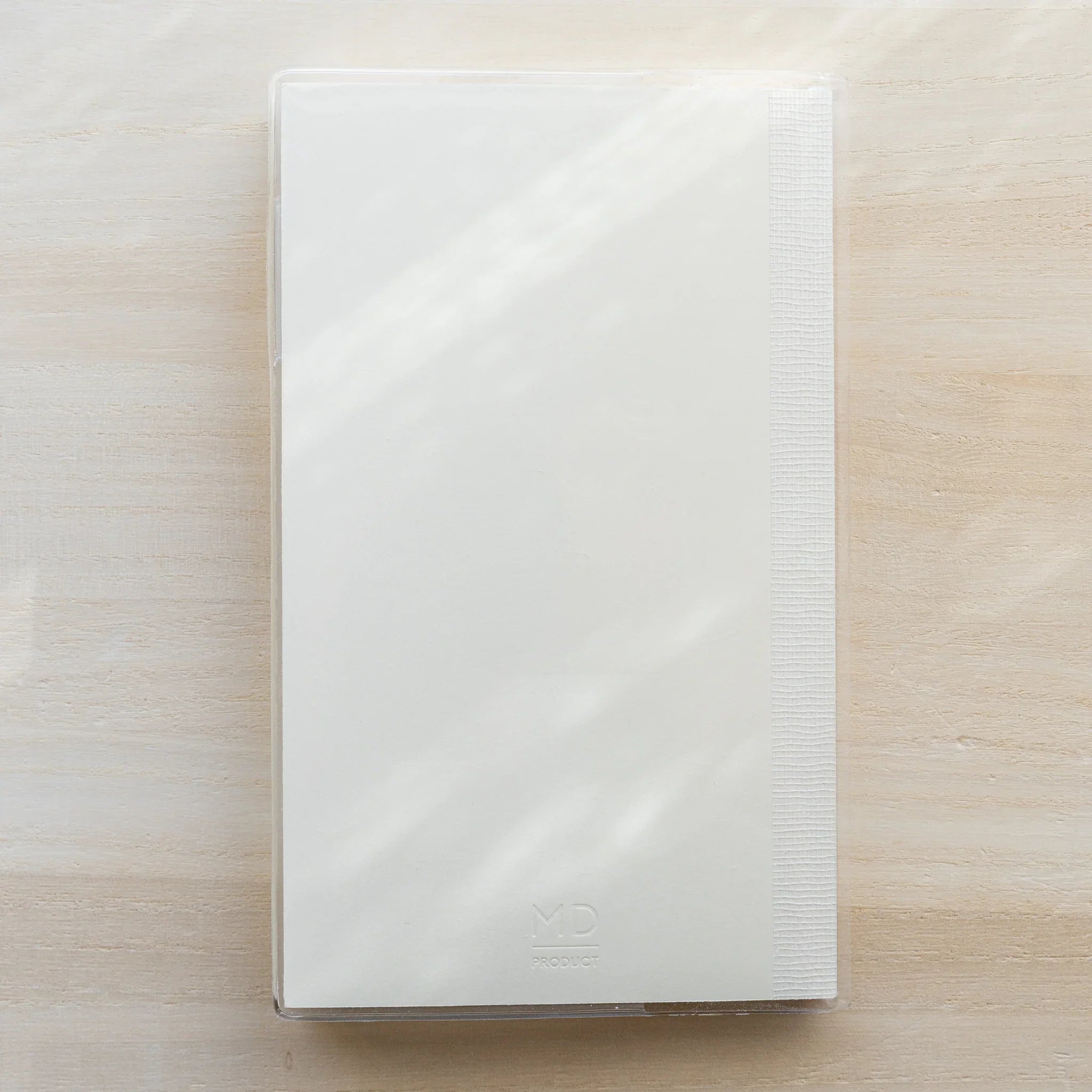 Midori MD Notebook B6 Slim in clear PVC cover, closed front view on wooden surface, white cover with ribbed spine on right