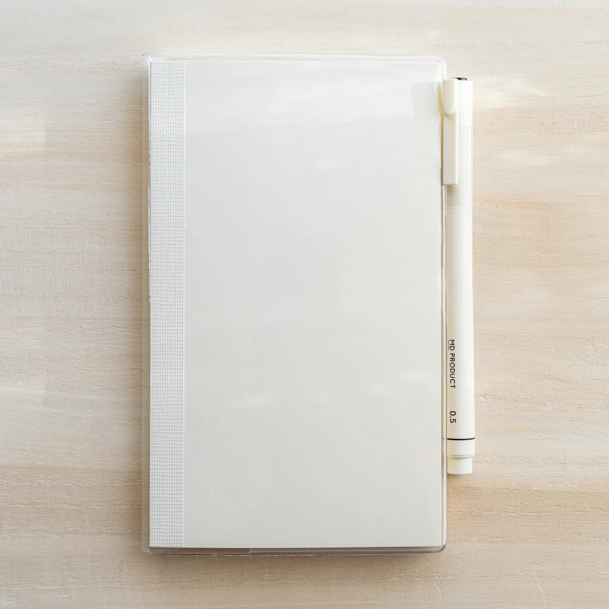 Midori MD Notebook A5 in clear PVC cover, flat lay on wooden surface with white MD PRODUCT pen resting to the right