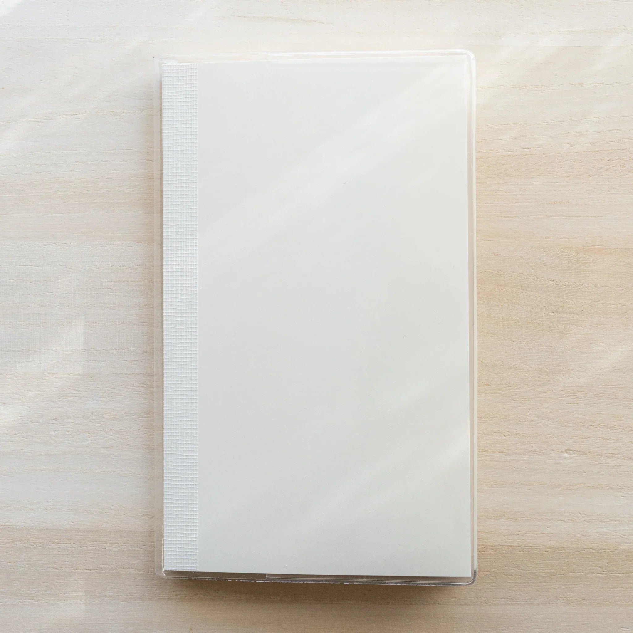 Midori MD Notebook A5 in clear PVC cover, flat lay top-down view on wooden surface, ribbed spine on left, plain white cover