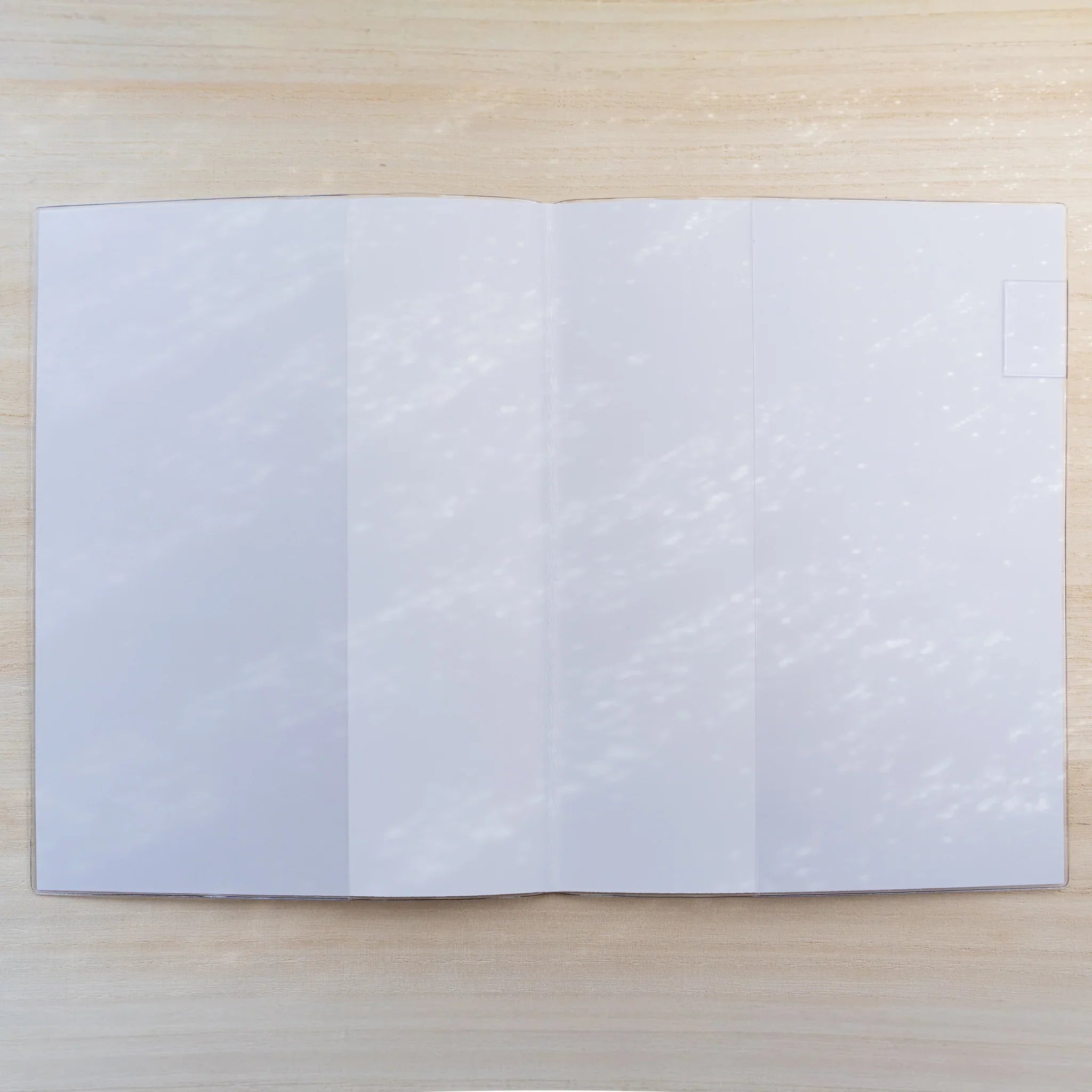 Midori MD Notebook Cover opened flat in clear PVC showing a blank white spread on a wood surface