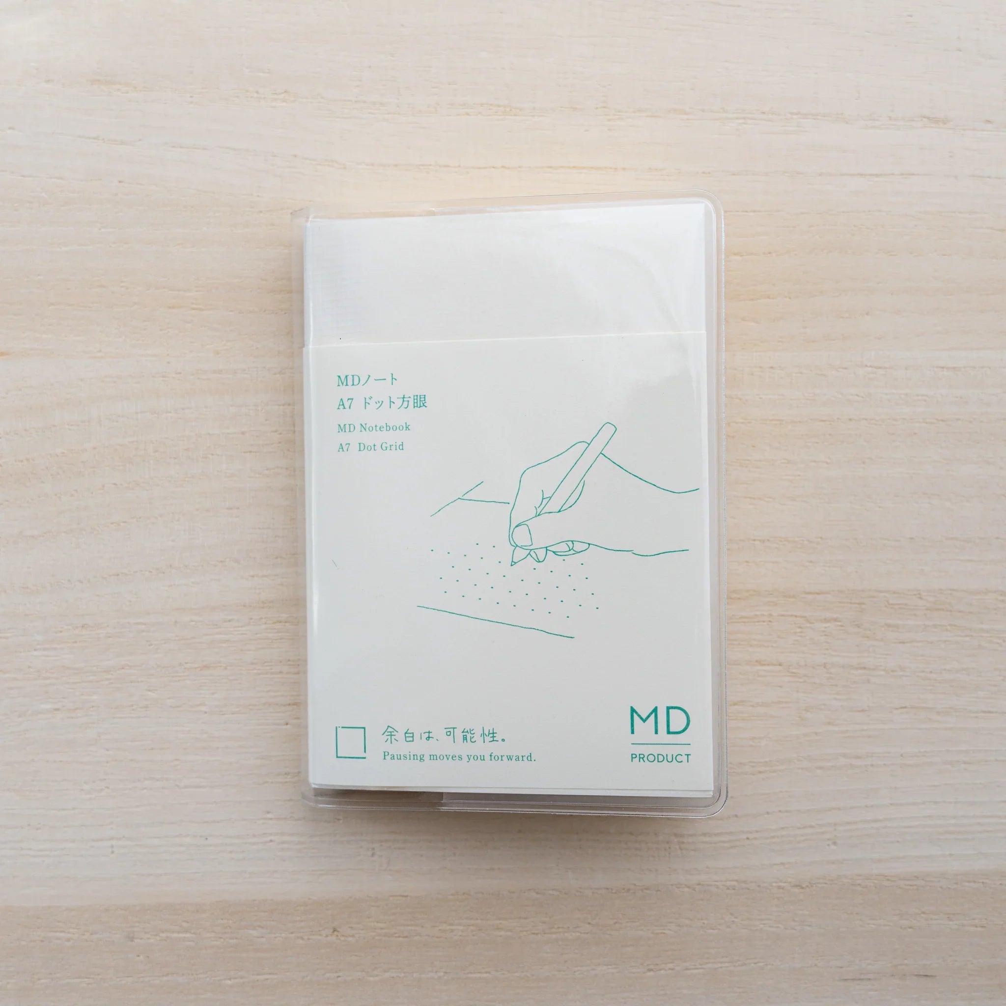 MD Notebook A7 Dot Grid in a clear vinyl cover with green hand-and-pen illustration and MD Product logo on wood