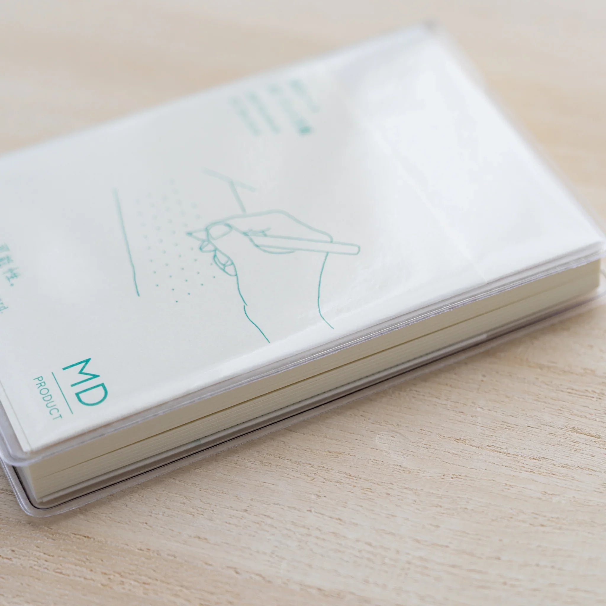 MD Notebook in a clear vinyl cover with green hand-and-pen illustration and MD Product logo, angled on a wood surface