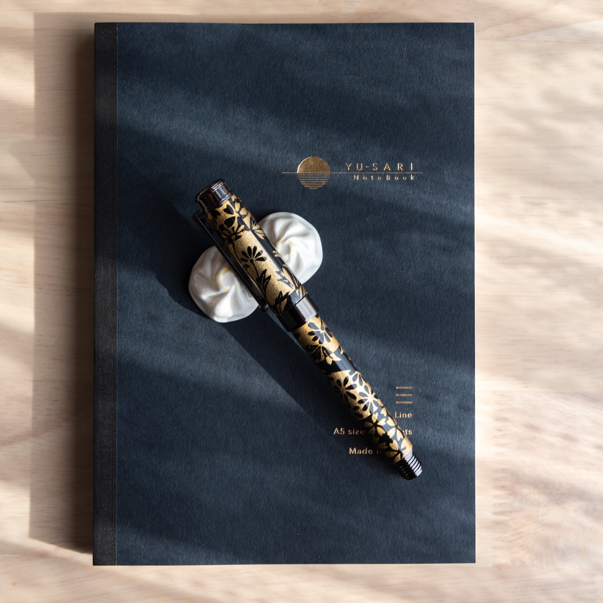 A Japanese fountain pen with Yuzen-patterned Mino washi rests on a white ceramic holder atop a black Yusari notebook, placed on a wooden desk.  